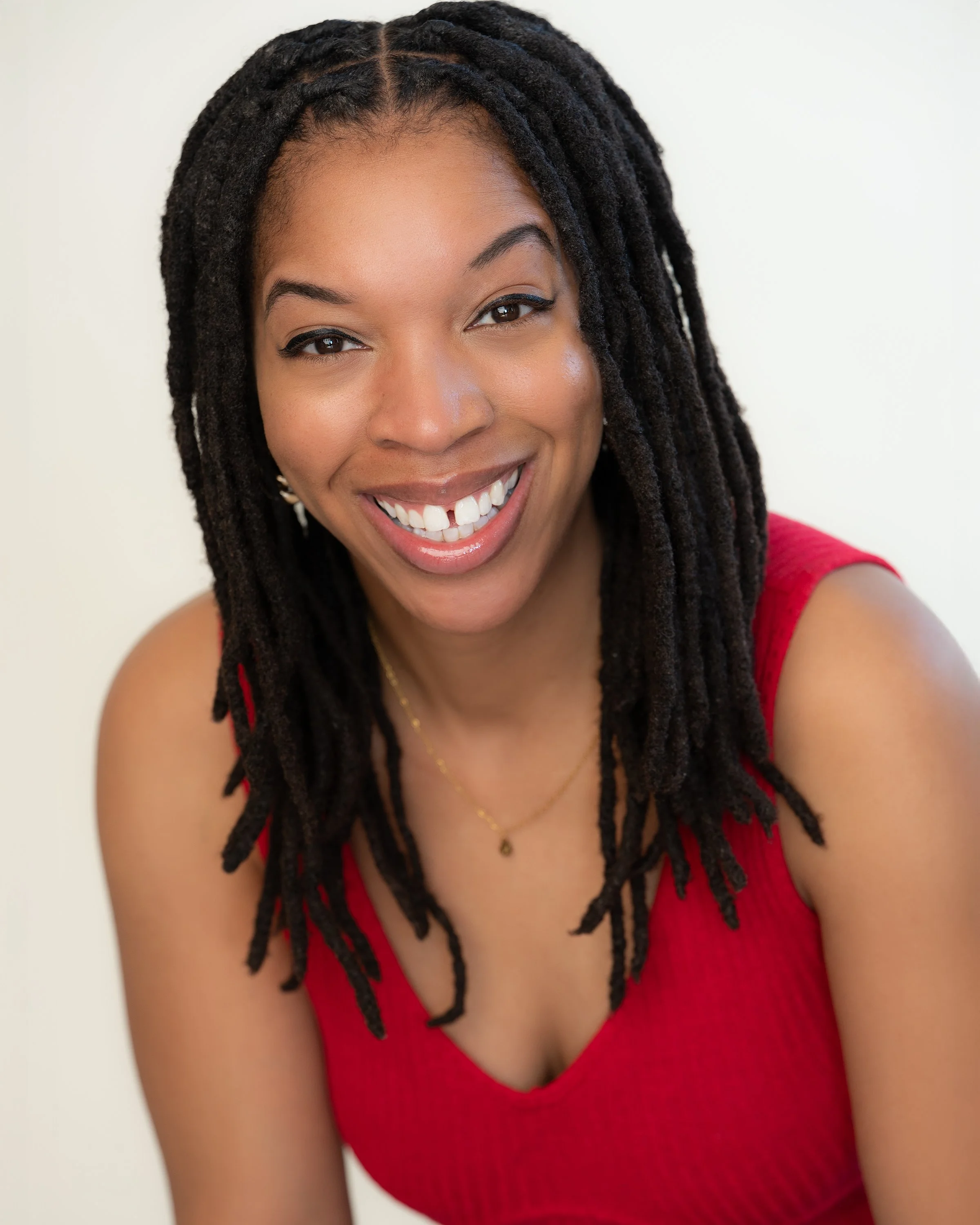 Close-up of a smiling woman with dark hair styled in dreadlocks, wearing a red sleeveless top, against a plain light background.