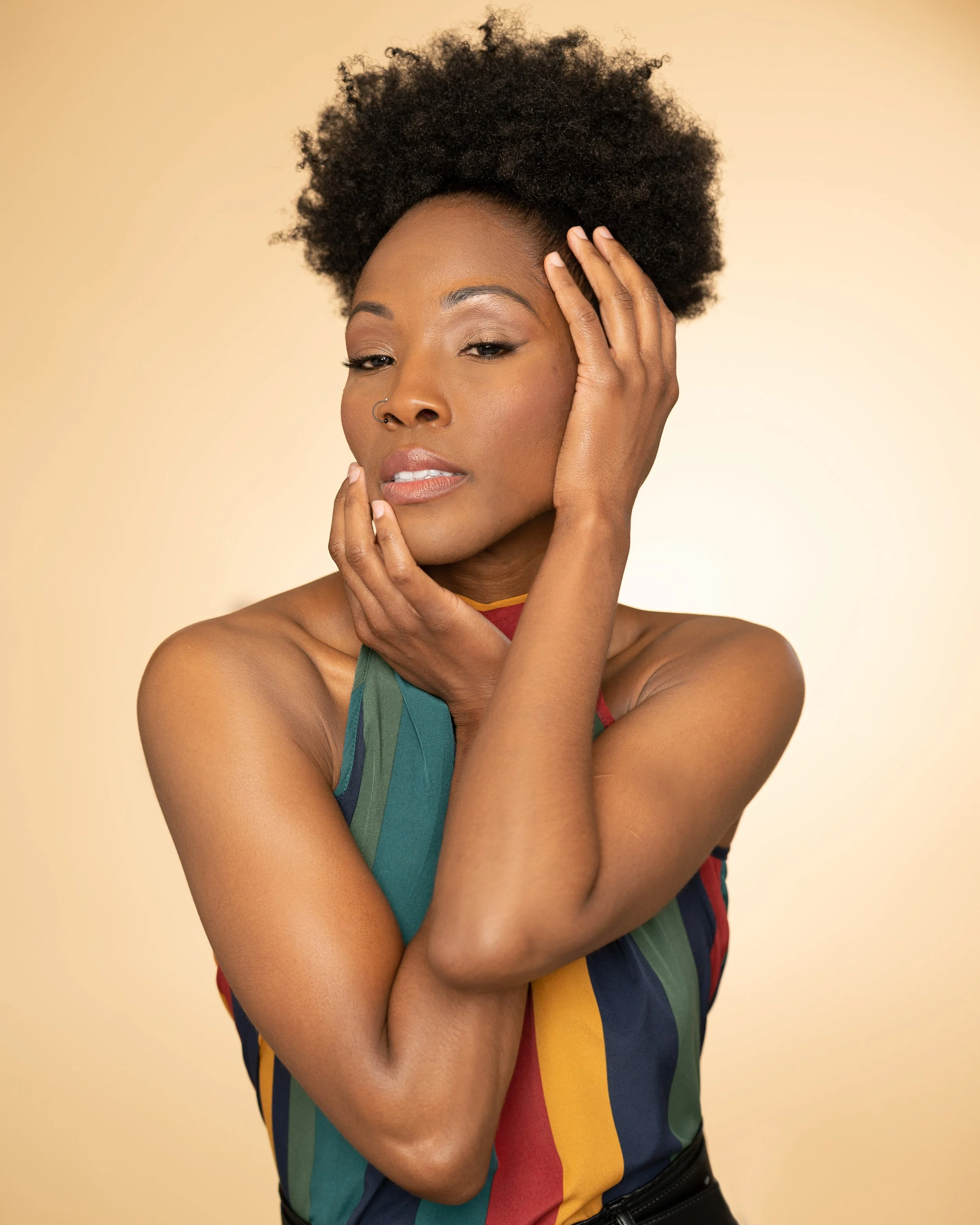 A woman with dark skin and curly short hair posing with one hand on her face and the other near her head, wearing a colorful striped top against a beige background.