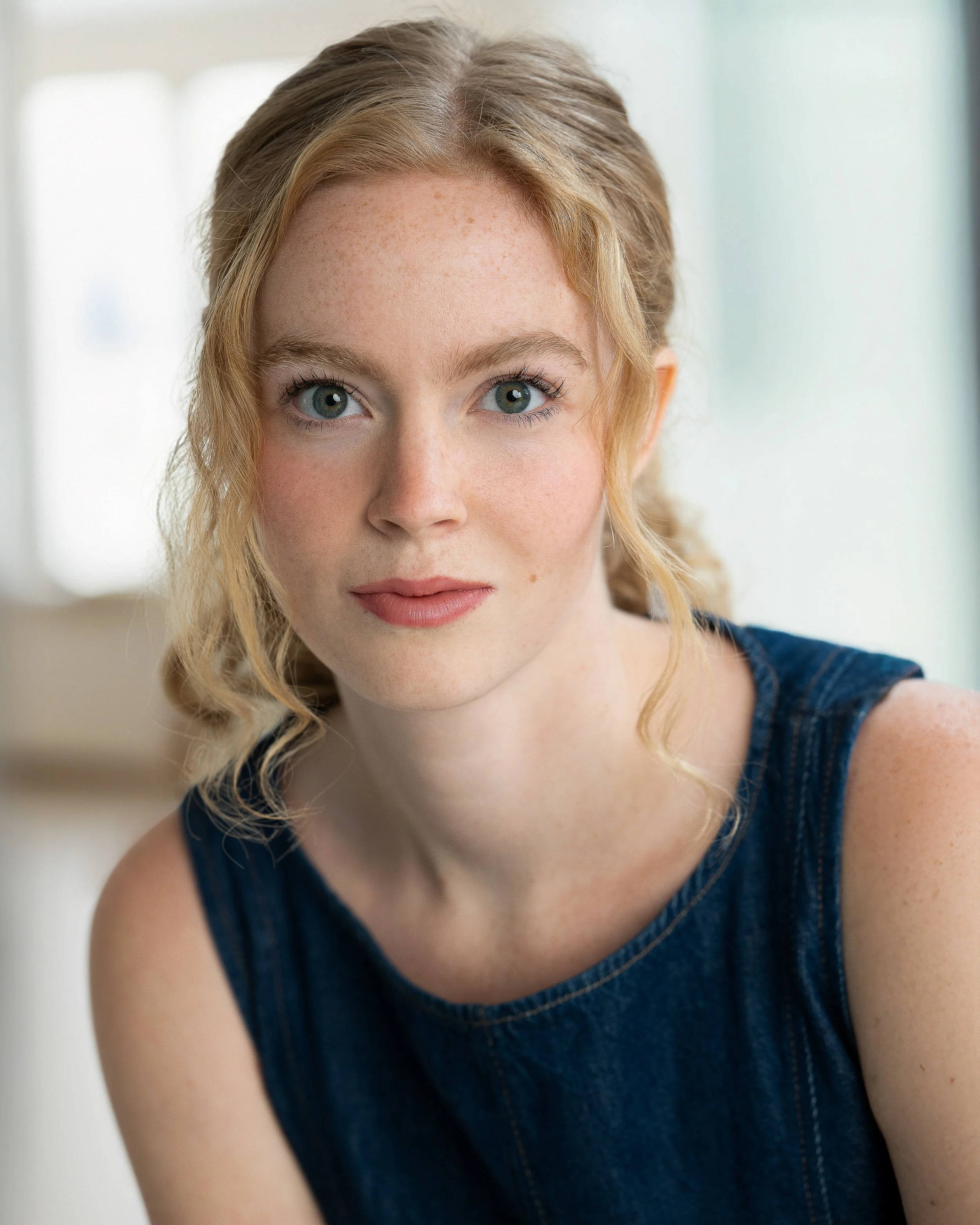 Close-up of a young woman with blonde curly hair, blue eyes, and fair skin, wearing a denim sleeveless top, indoors with soft natural light.