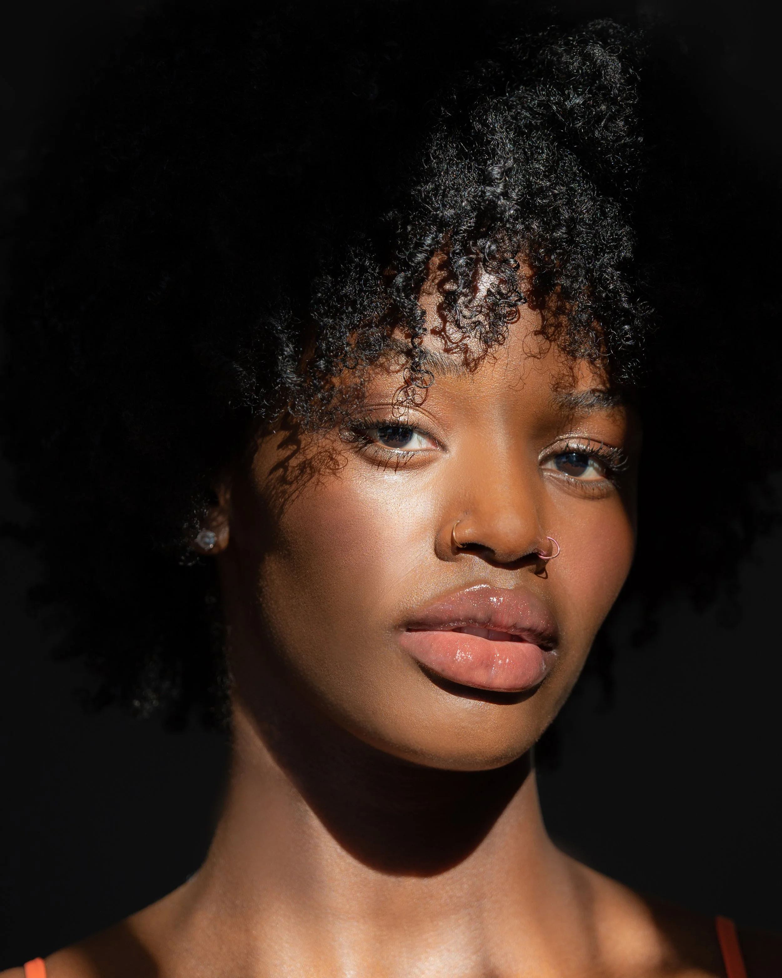 Close-up of a person with dark curly hair covering part of their face, focus on their eyes and lips, with natural makeup and a warm skin tone.