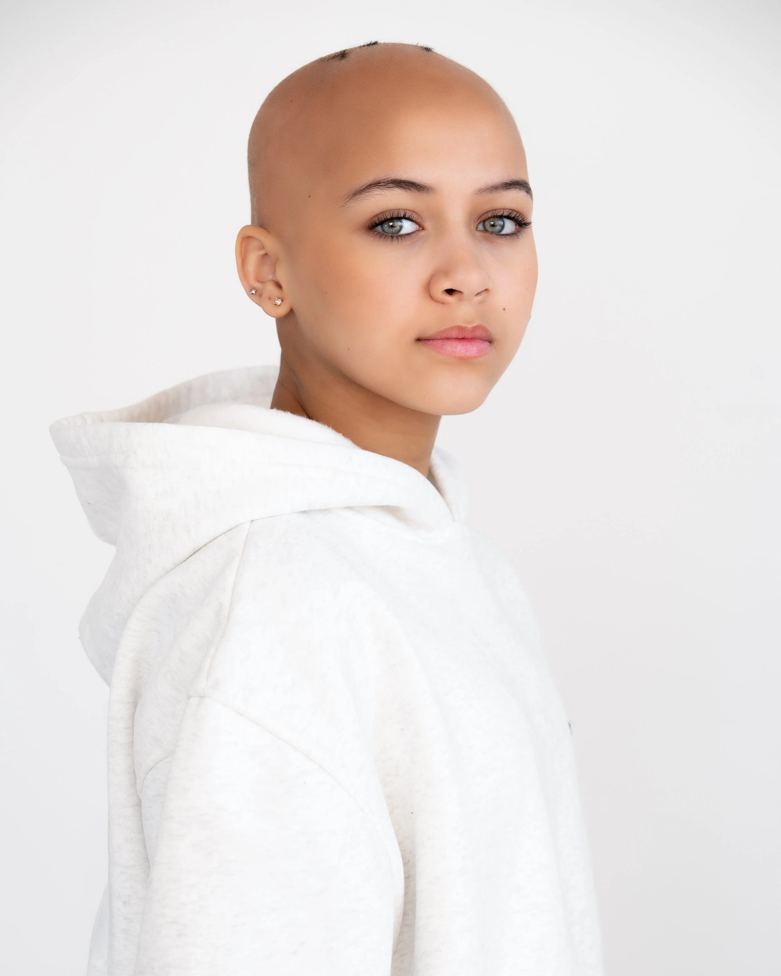 A young woman with a shaved head and blue eyes, wearing earrings and a white hoodie, looking directly at the camera against a plain white background.