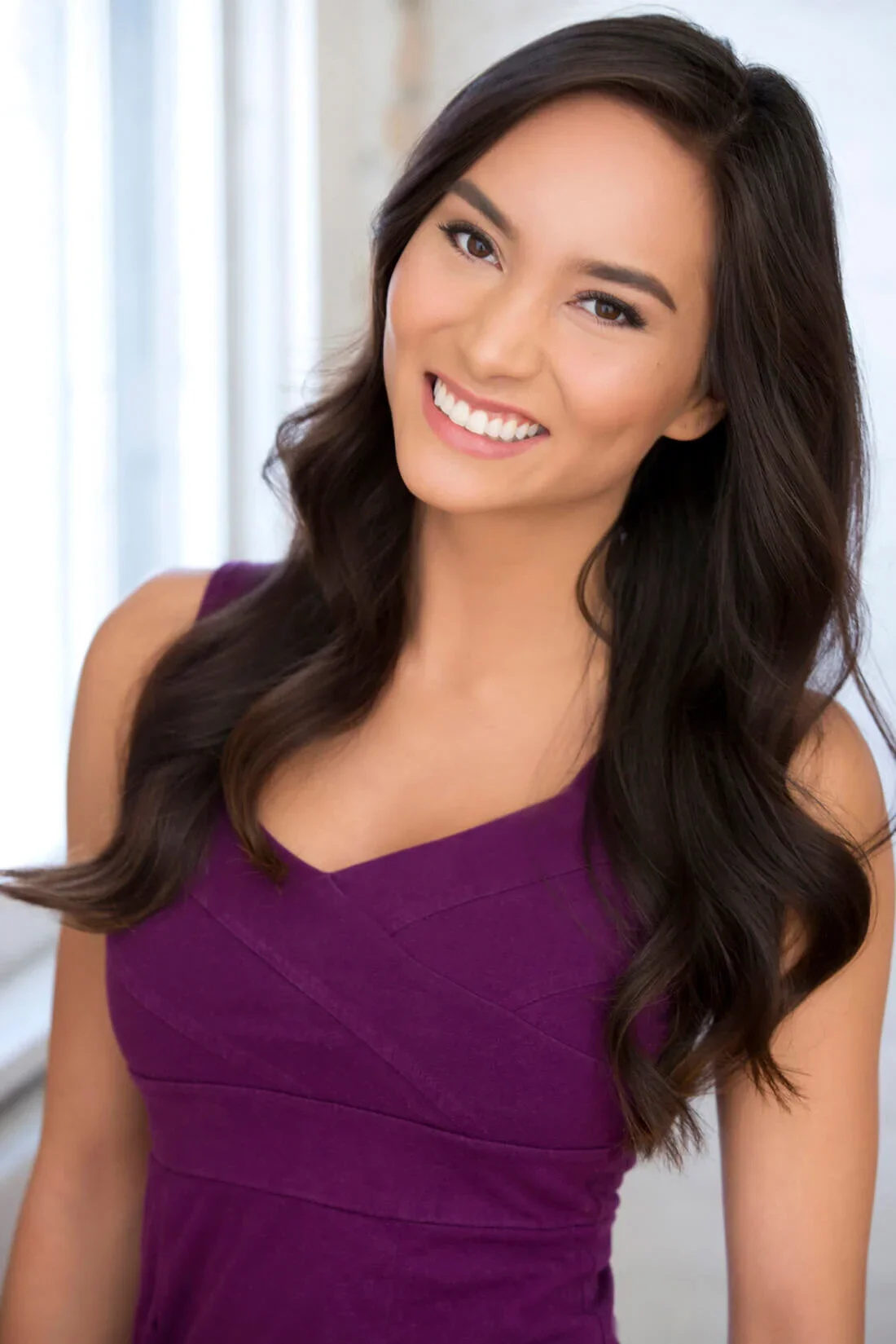 A woman smiling with long dark hair, wearing a sleeveless purple dress, standing in a bright room with a blurred background.