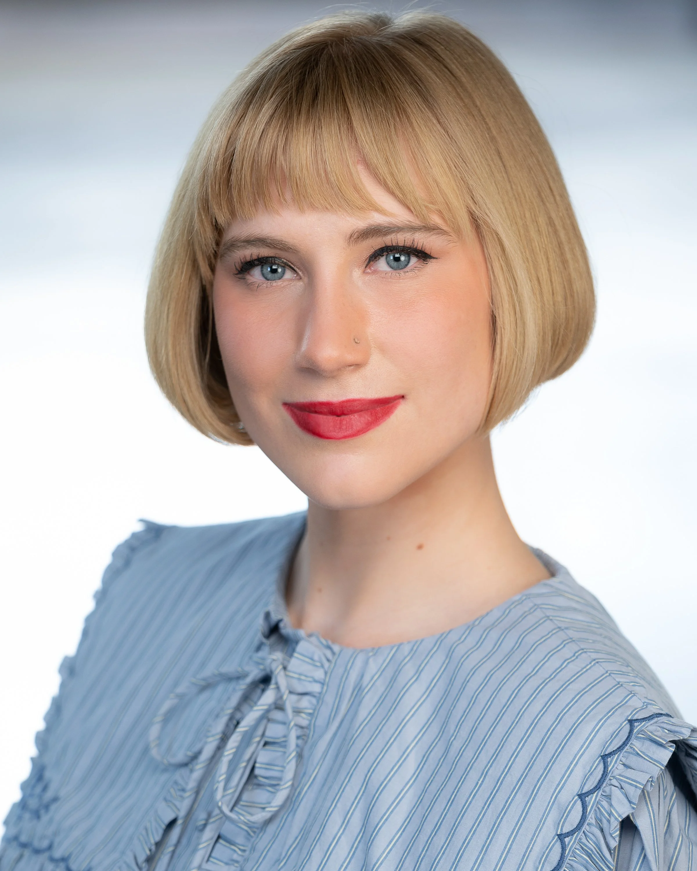A woman with short blonde hair, blue eyes, red lipstick, and a nose piercing, wearing a light blue striped shirt with ruffled sleeves, looking at the camera with a slight smile.