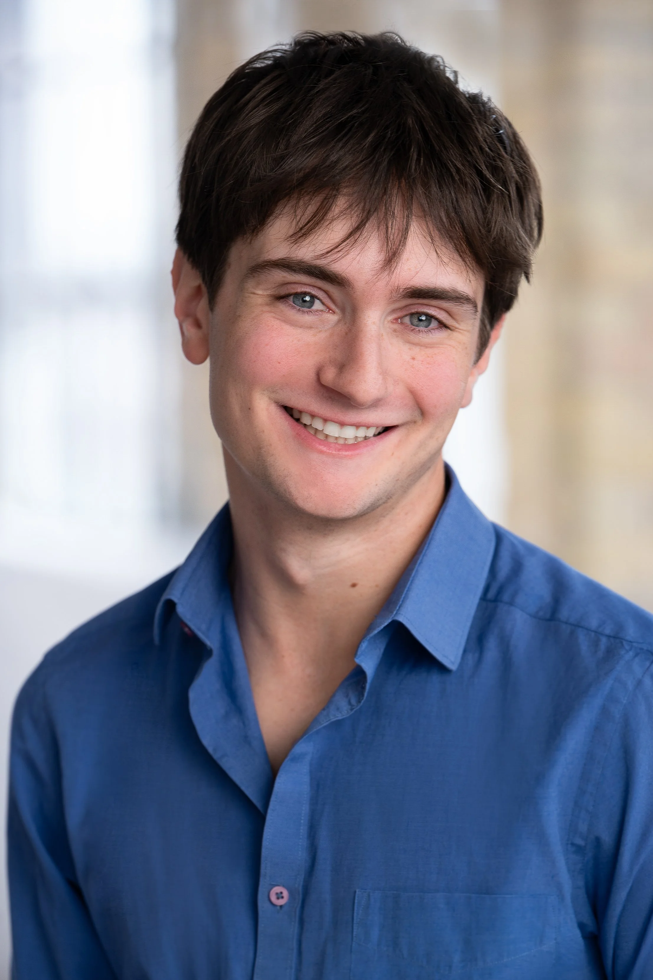 Close-up portrait of a young man with dark brown hair, light blue eyes, and fair skin, smiling and wearing a blue button-up shirt.