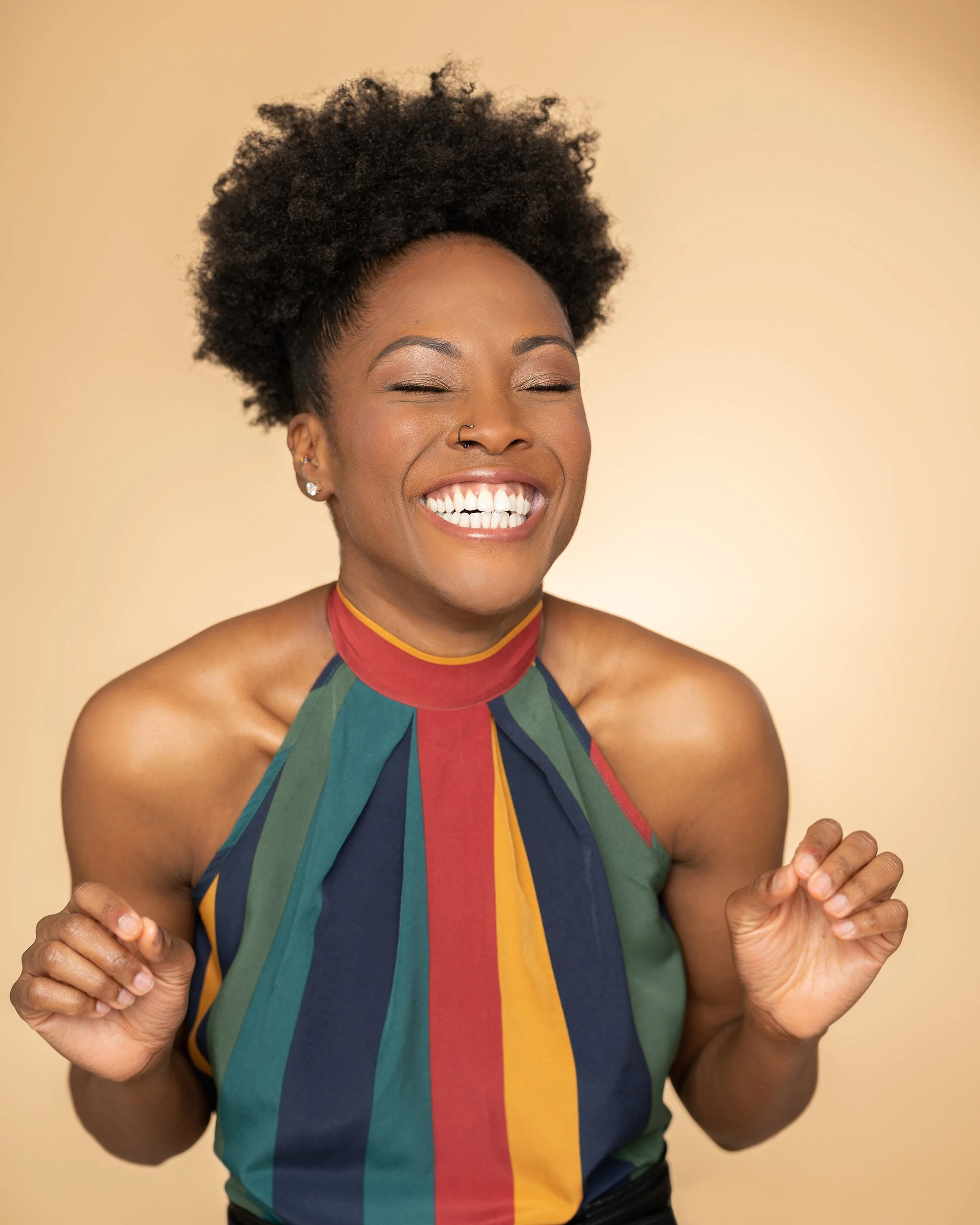 A joyful Black woman with natural hair, smiling brightly with eyes closed, wearing a colorful halter top with vertical stripes in red, blue, green, yellow, and orange, standing against a plain beige background.