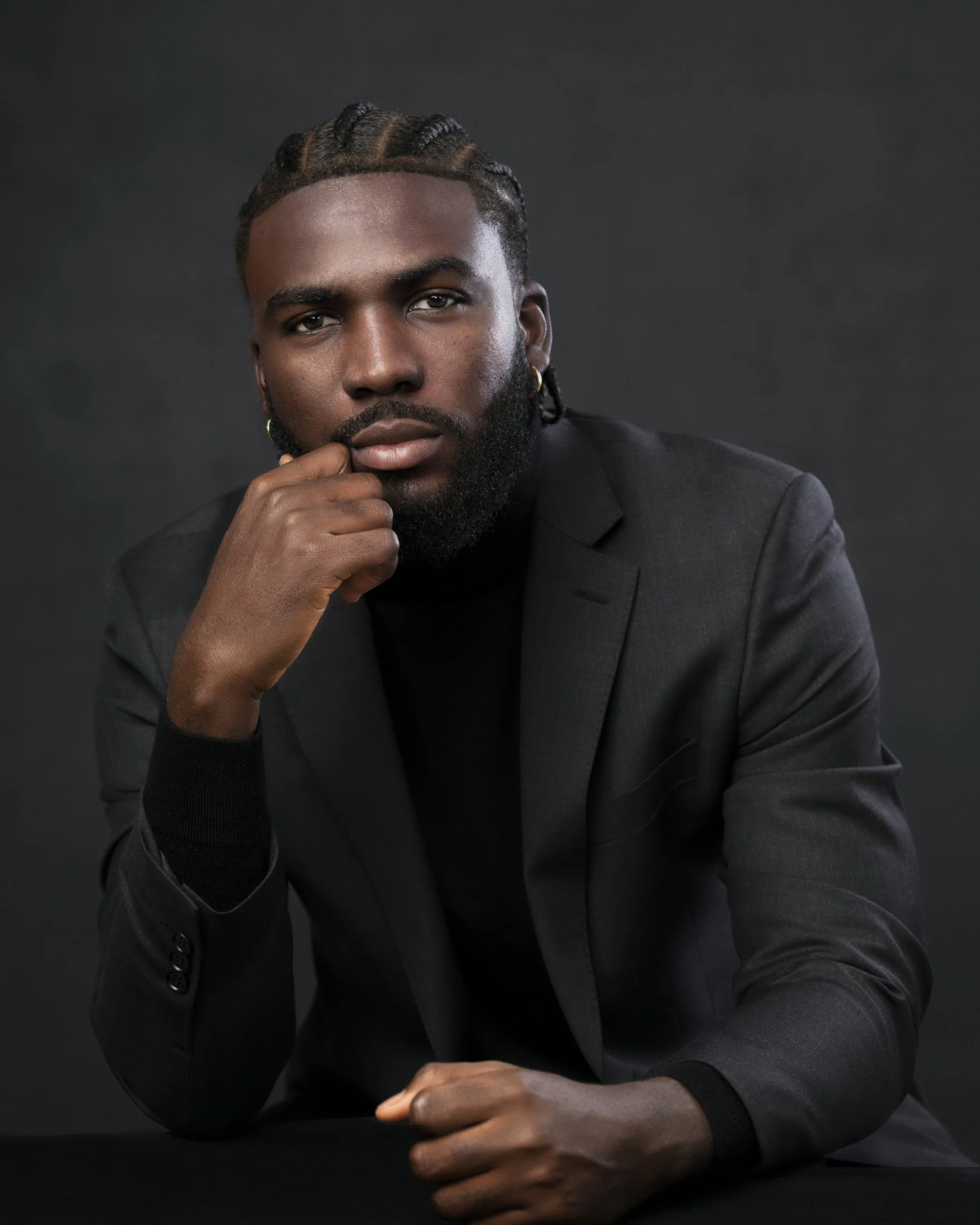 A Black man with styled hair, a beard, and earrings, wearing a black suit, posing against a dark background.