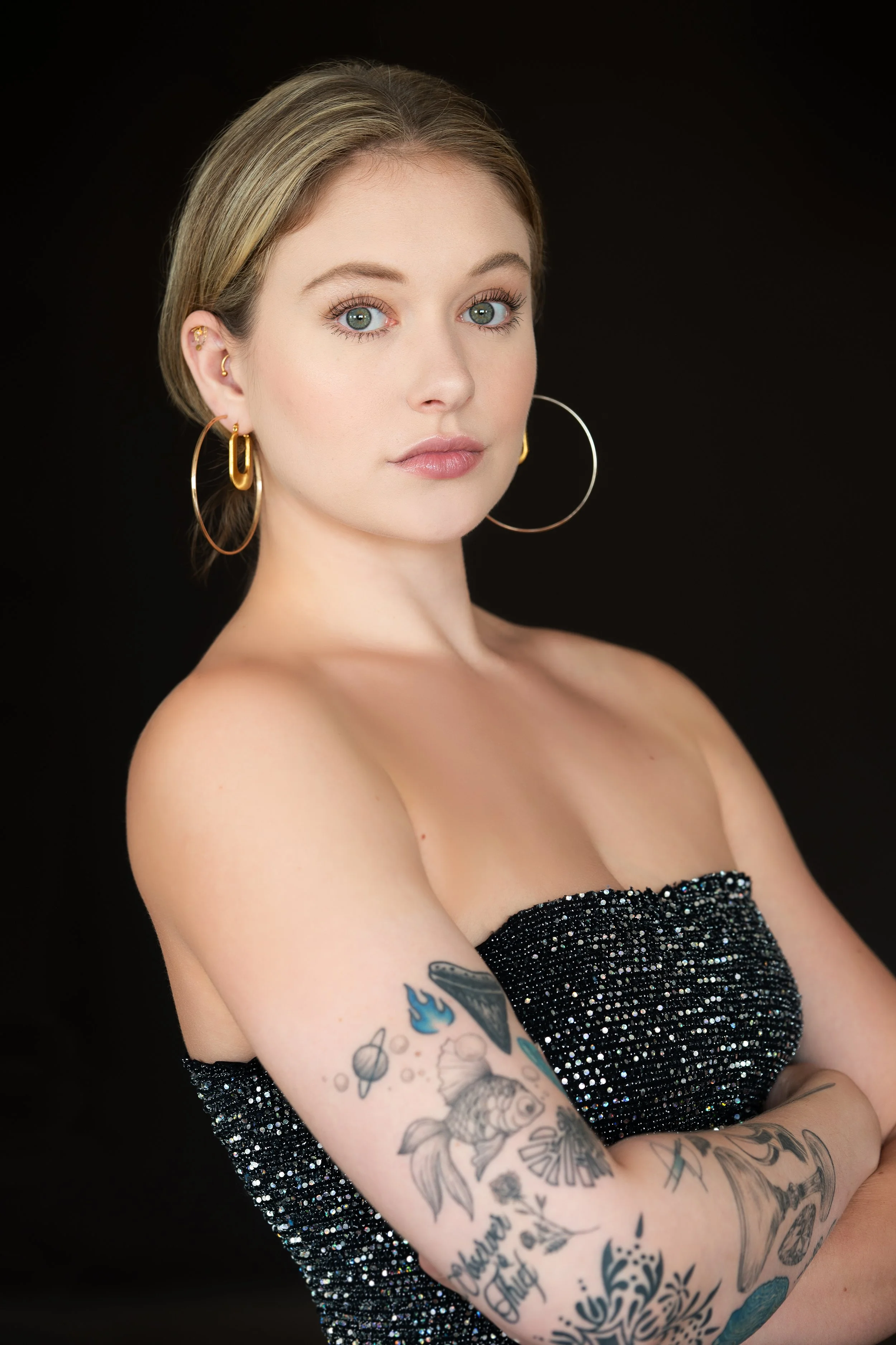 A young woman with blonde hair, blue eyes, and light skin posing against a black background. She is wearing large gold hoop earrings and a strapless black dress embellished with sparkles. She has tattoos on her arm, including a fish, planets, and var