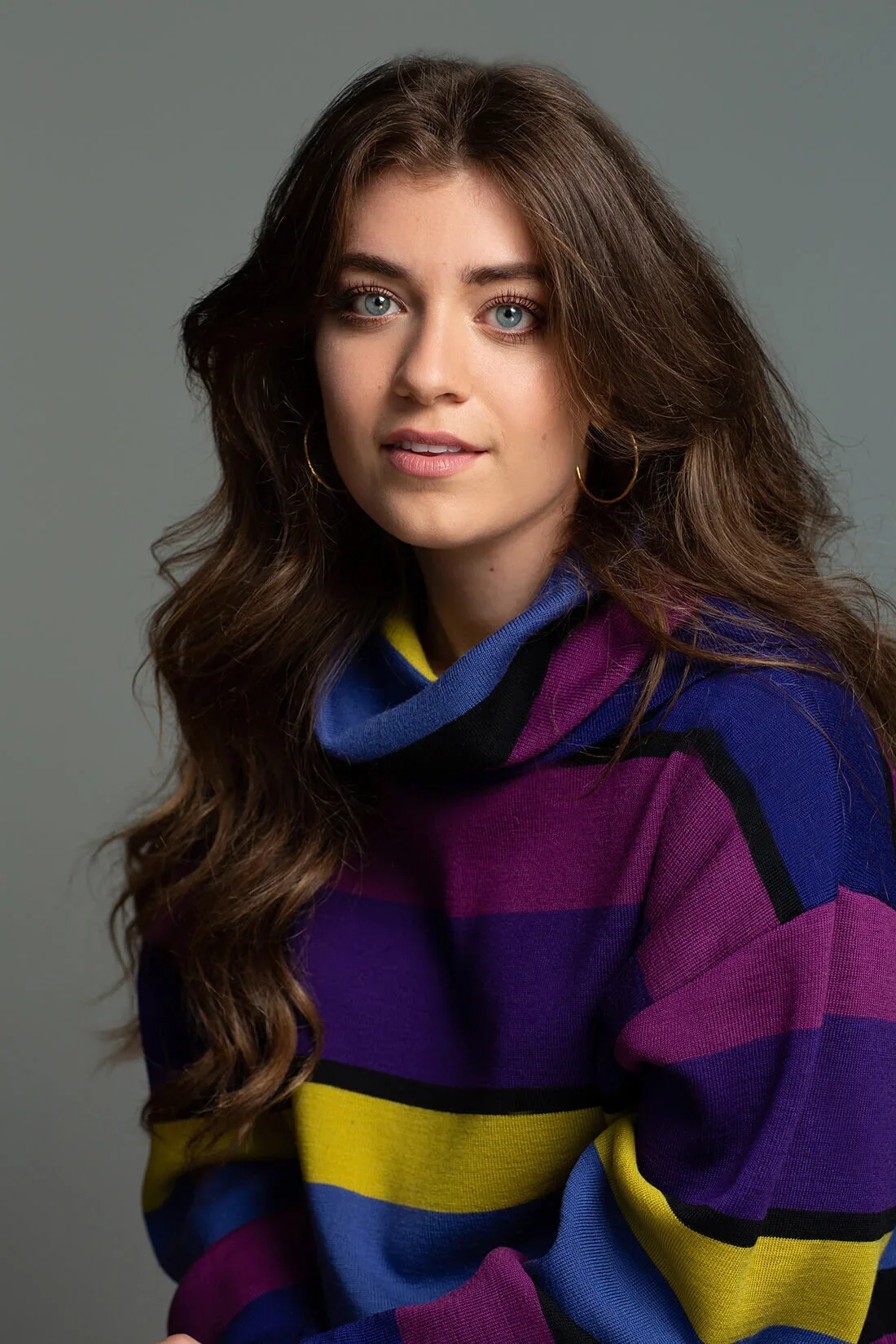 A young woman with long, wavy brown hair and blue eyes, wearing a color-blocked sweater in purple, yellow, blue, and black, and gold hoop earrings, posing against a plain gray background.