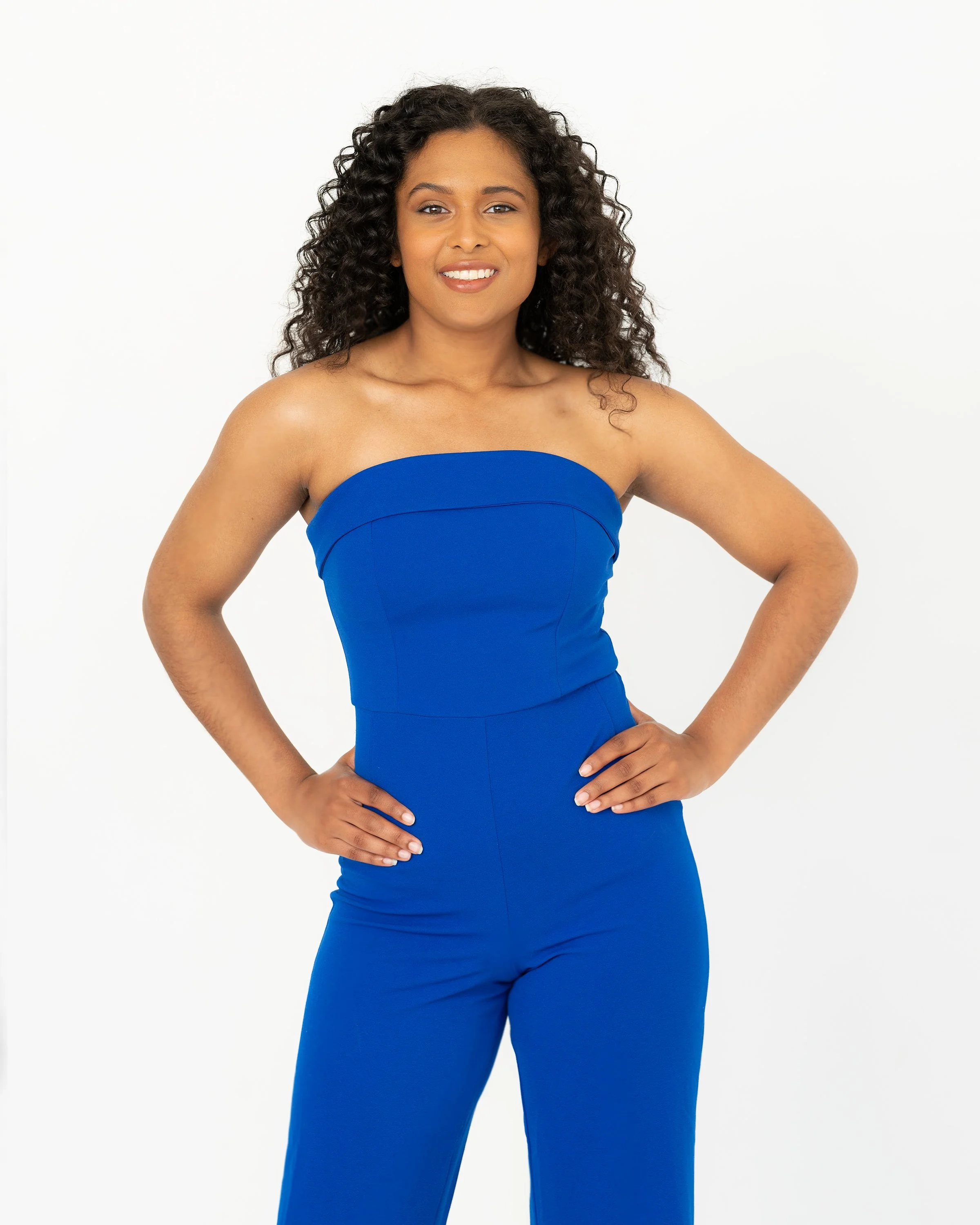 A woman with dark curly hair, wearing a strapless royal blue dress, stands with her hands on her hips and a confident smile against a plain white background.