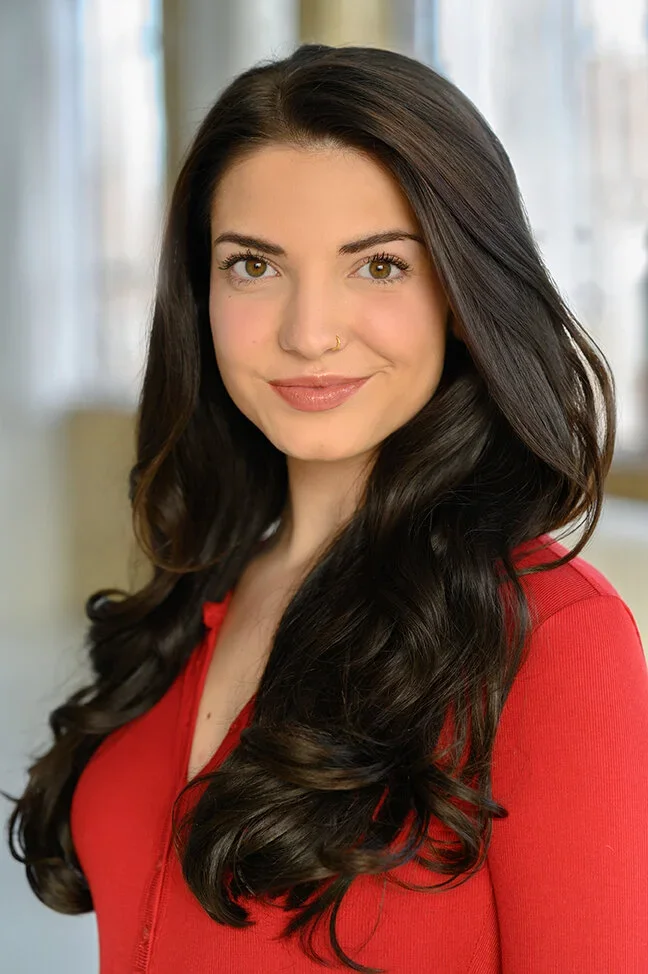 A young woman with long, dark wavy hair wearing a red top, smiling indoors with a blurred background of curtains and a window.