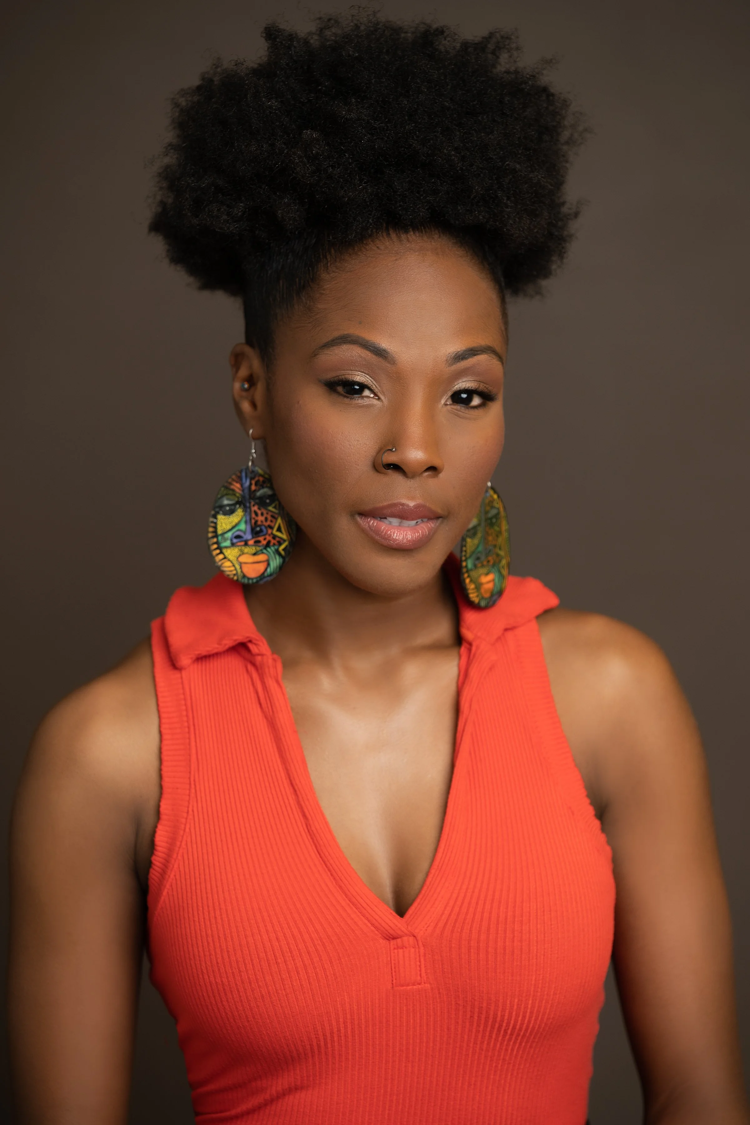 A woman with natural curly hair, wearing colorful earrings with African patterns and a red sleeveless top, posing against a plain dark background.