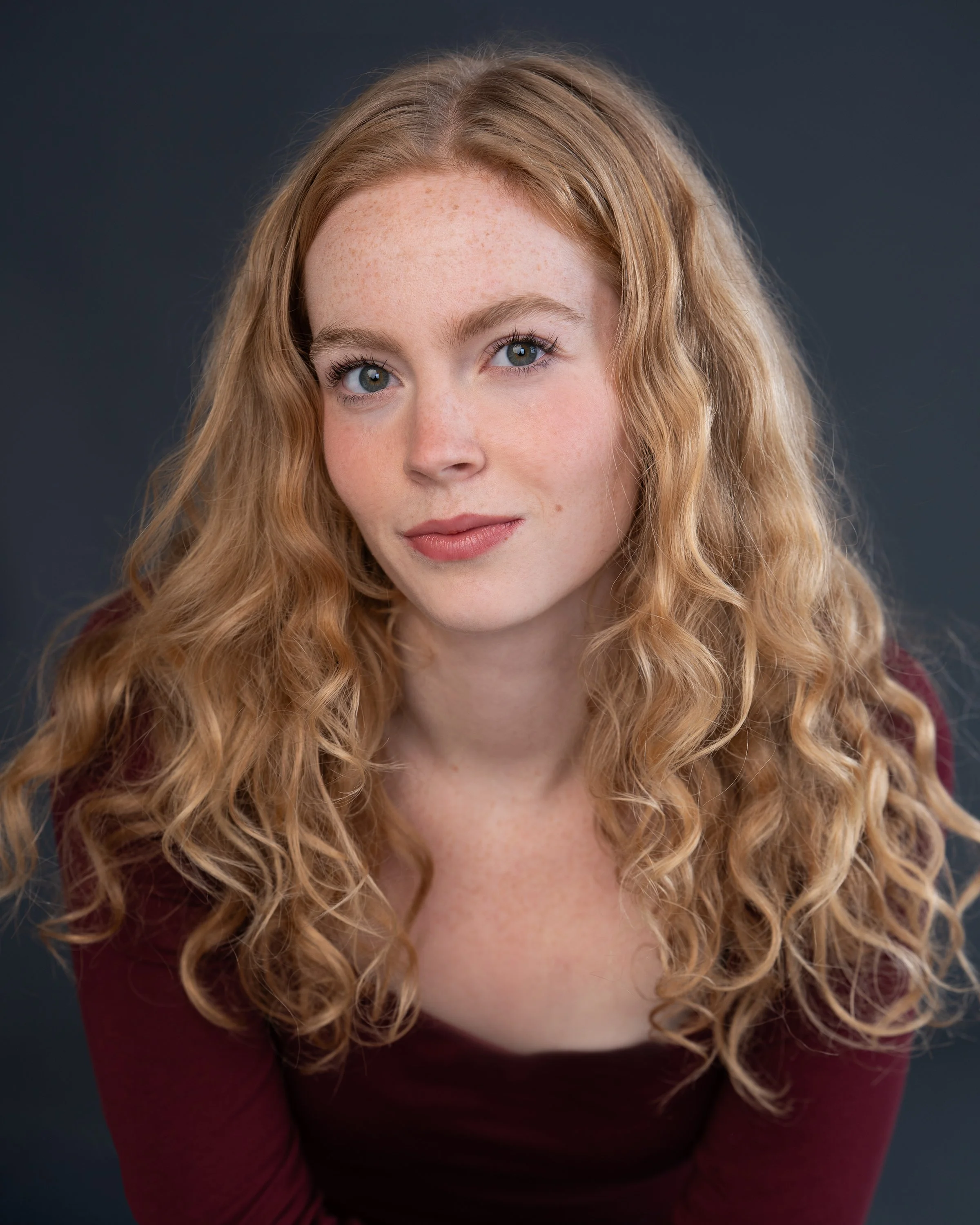 Close-up of a woman with wavy red hair, fair skin, and blue eyes, looking into the camera with a slight smile, wearing a maroon top against a dark background.