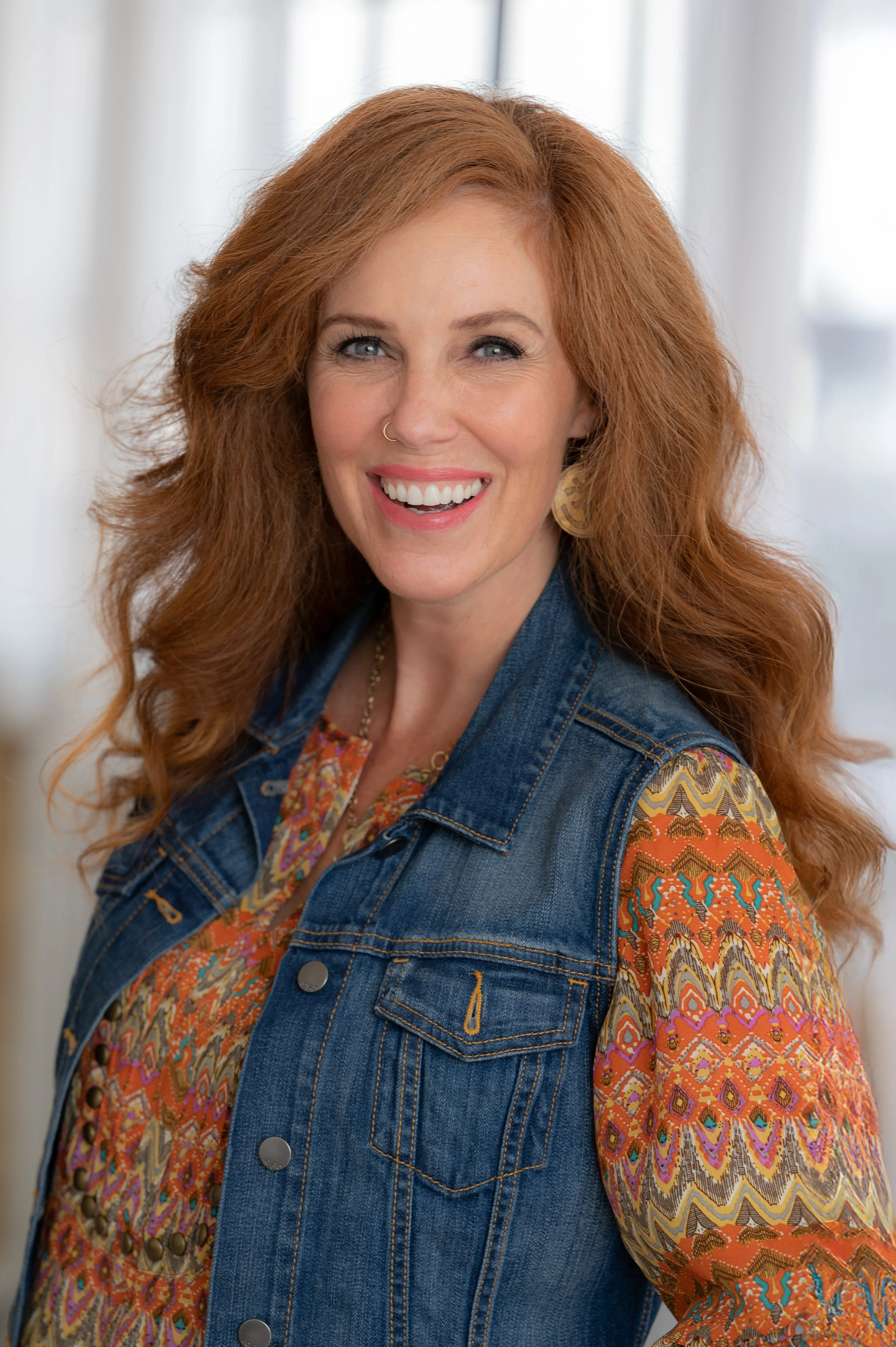 A woman with red hair, wearing a denim vest over a colorful patterned blouse, smiling at the camera.