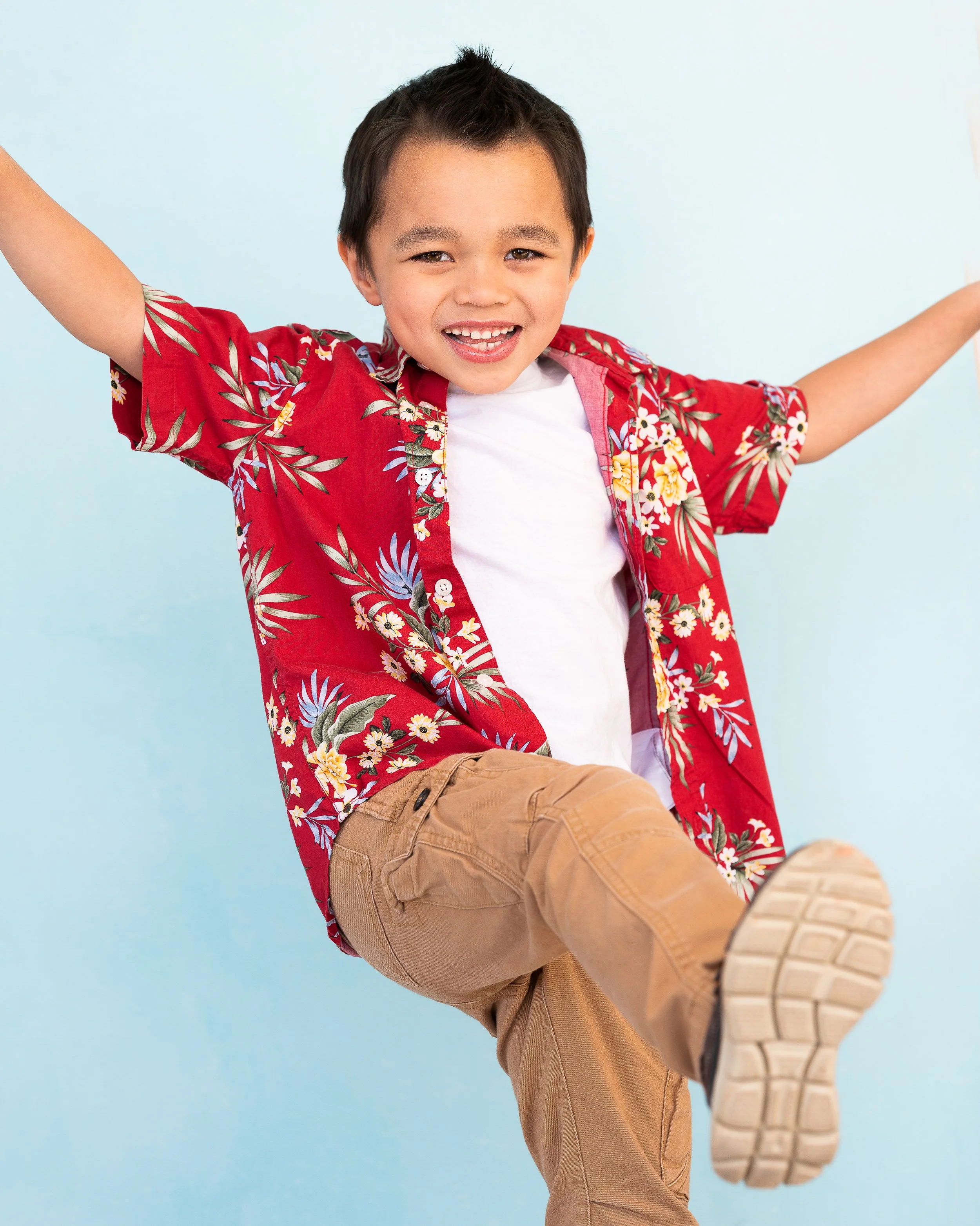 A young boy with dark hair smiling and raising his arms, wearing a red floral patterned shirt, white T-shirt, and tan pants against a light blue background.