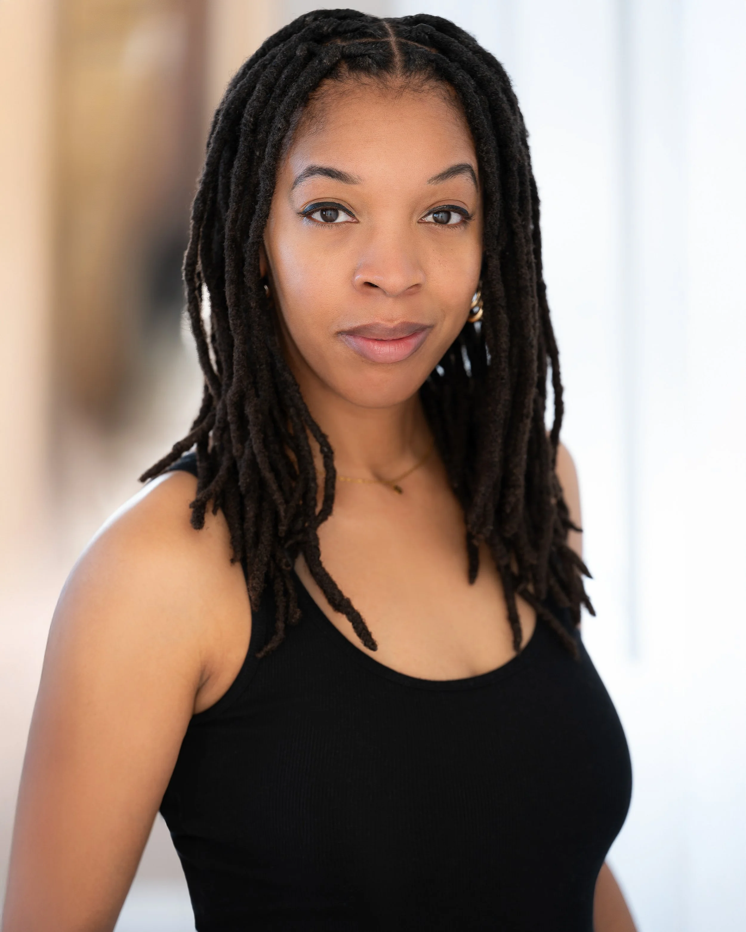 A young woman with medium brown skin and long black dreadlocks styled over her shoulders, wearing a black sleeveless top, standing indoors with a neutral background.