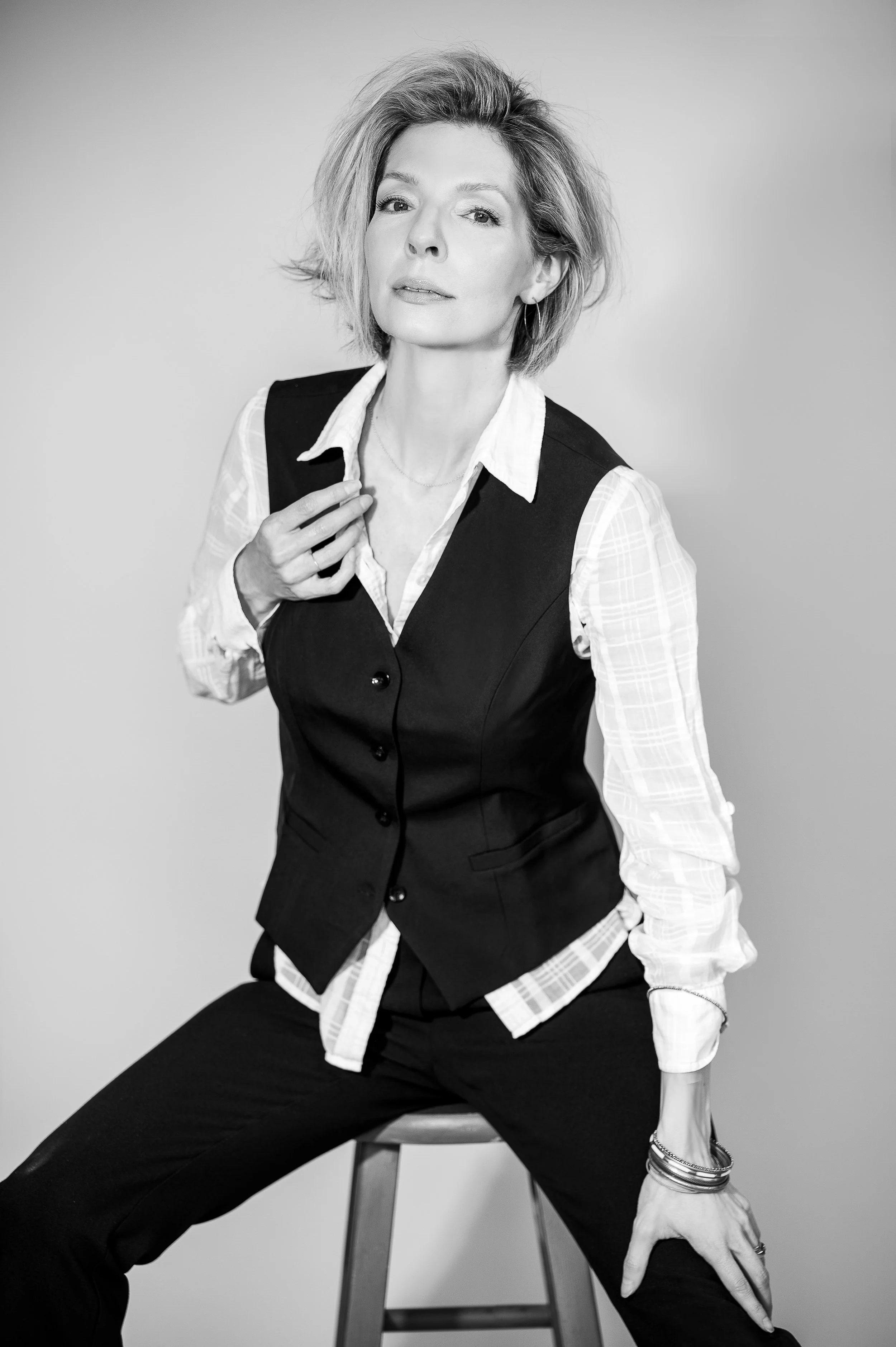 Black and white photo of a woman with short, wavy hair sitting on a stool, wearing a white shirt under a dark vest, with one hand resting on her lap and the other on her chest, looking confidently at the camera.