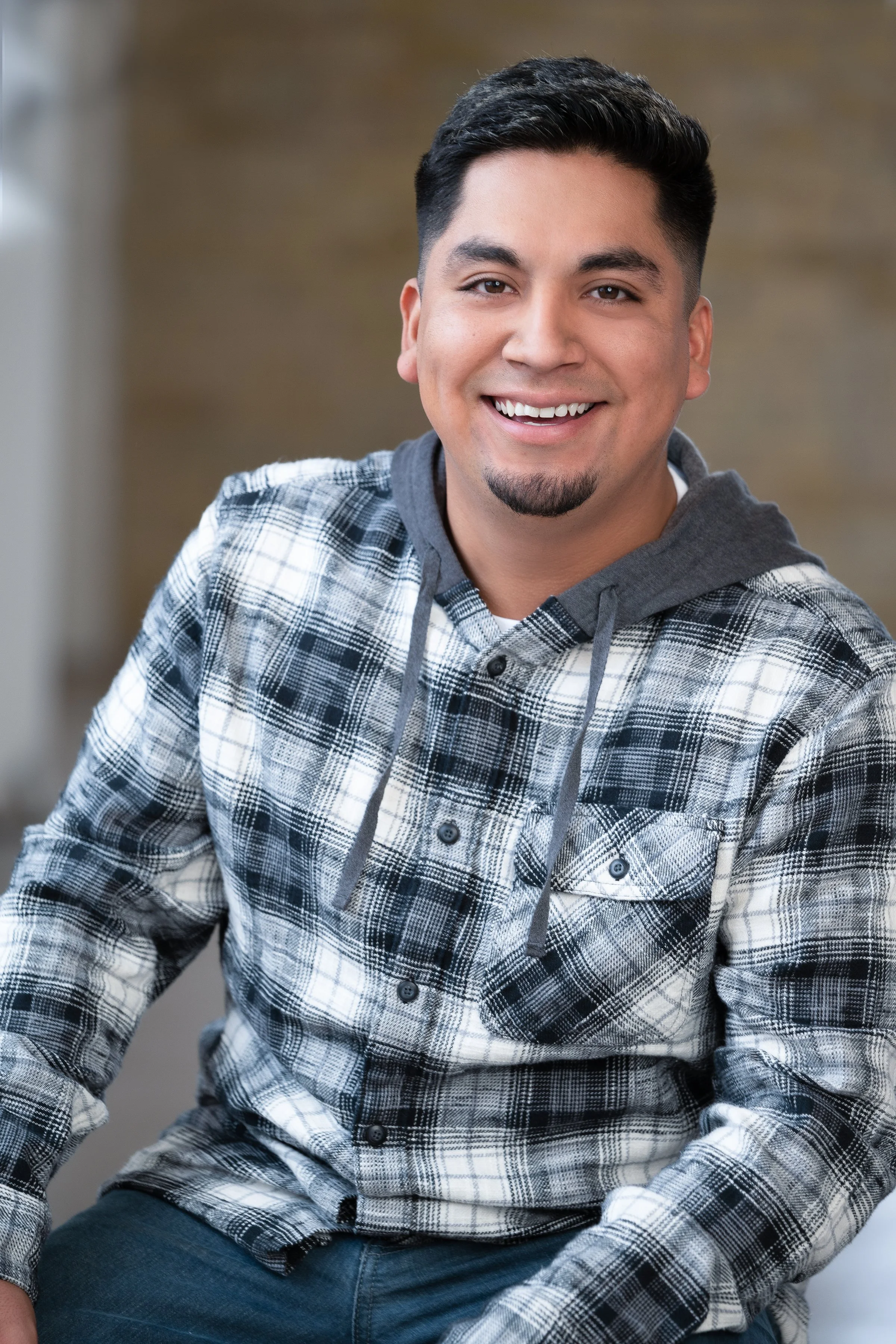 A smiling young man with short black hair, a goatee, wearing a plaid shirt with a hoodie underneath, sitting indoors.