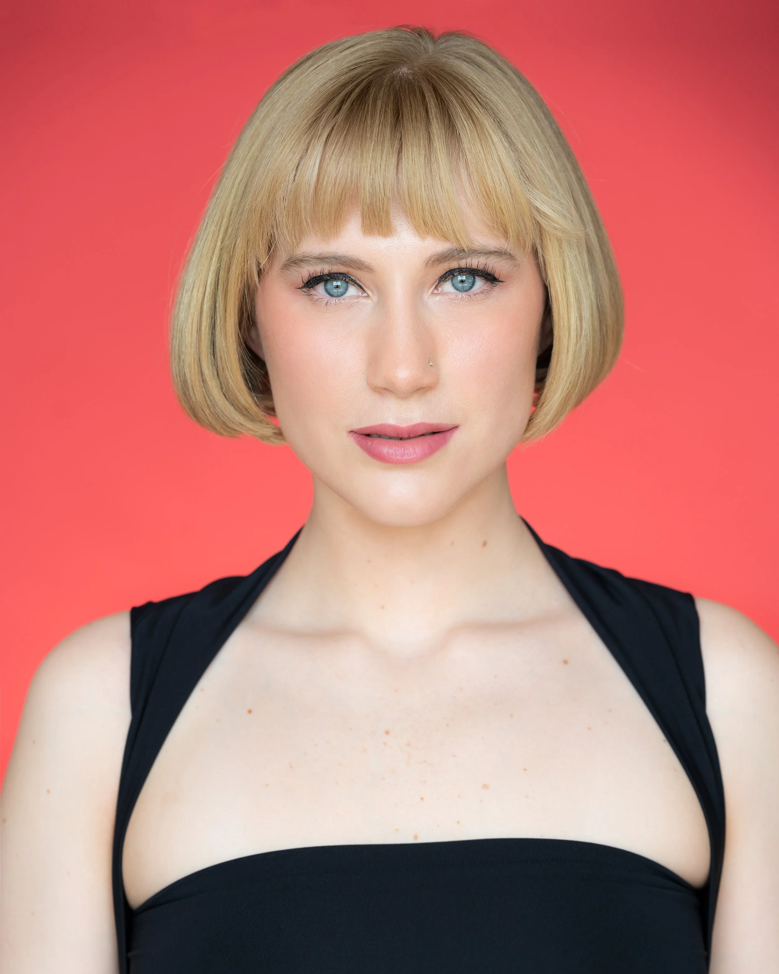 Close-up portrait of a woman with blond hair in a short bob, blue eyes, wearing a black top, against a pink background.