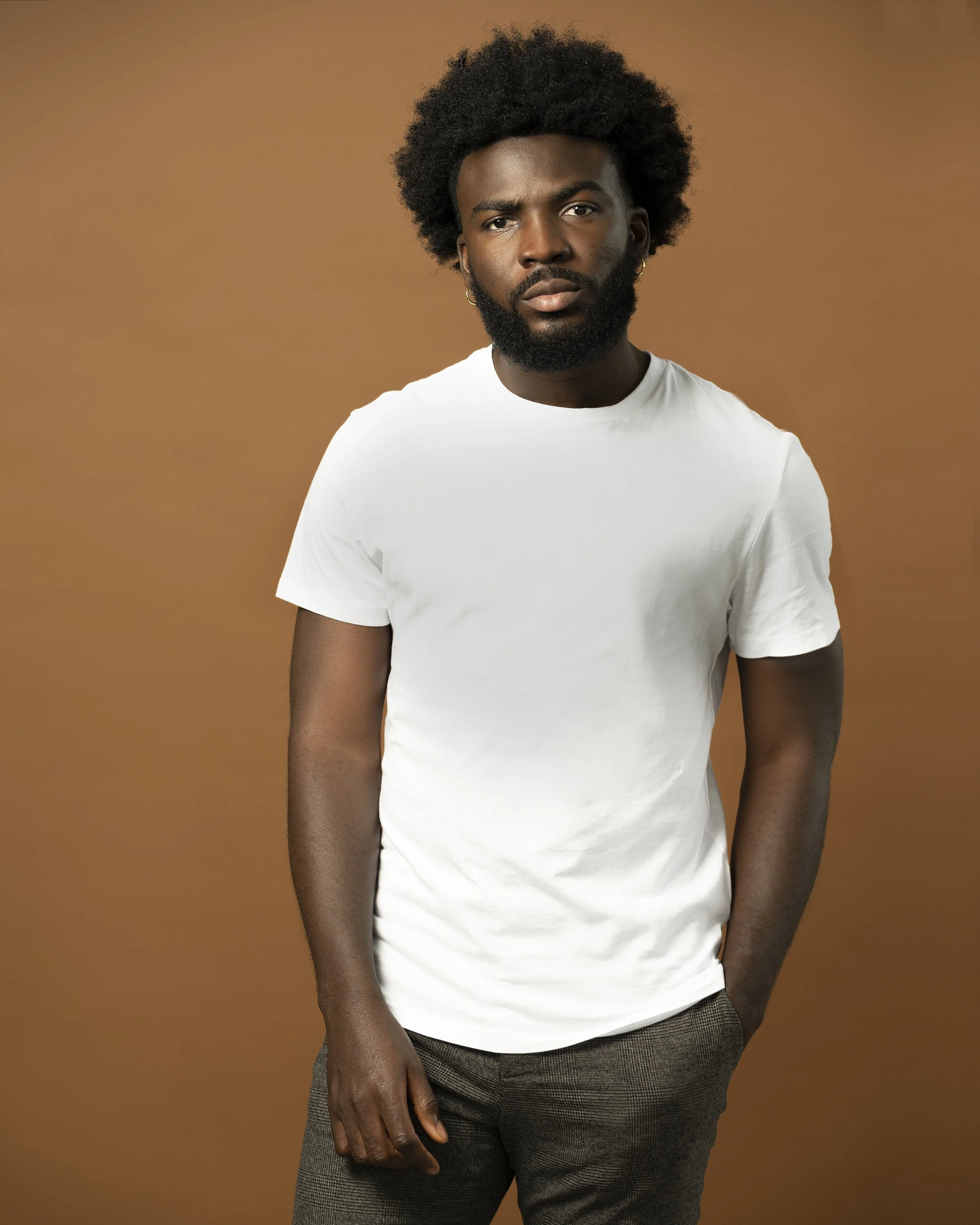 Portrait of a man with curly black hair and a beard, wearing a white T-shirt and gray plaid pants, standing against a brown background.