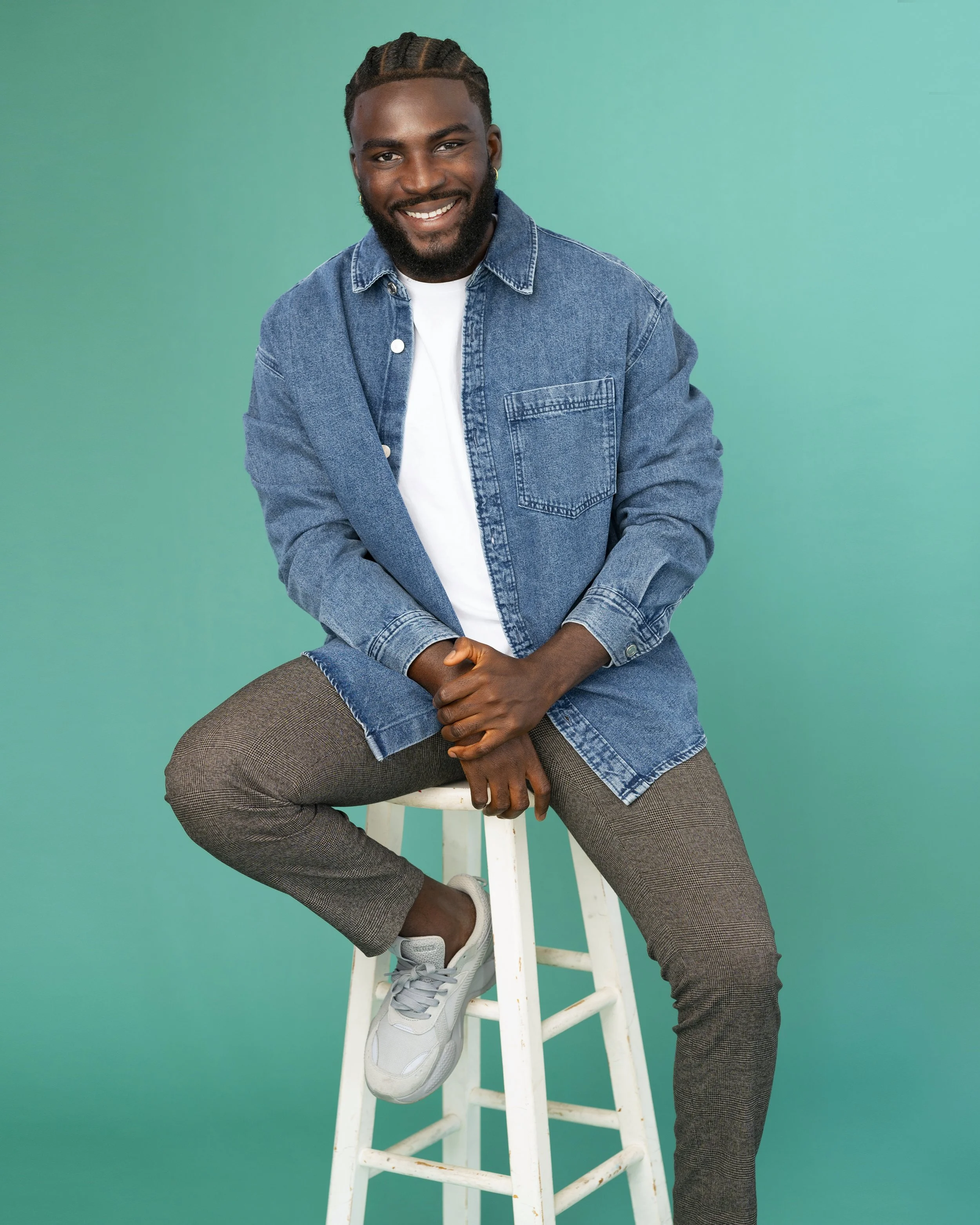 A young man with braids, a beard, and earrings, smiling while sitting on a white stool against a teal background. He is wearing a denim jacket, white t-shirt, plaid pants, and white sneakers.