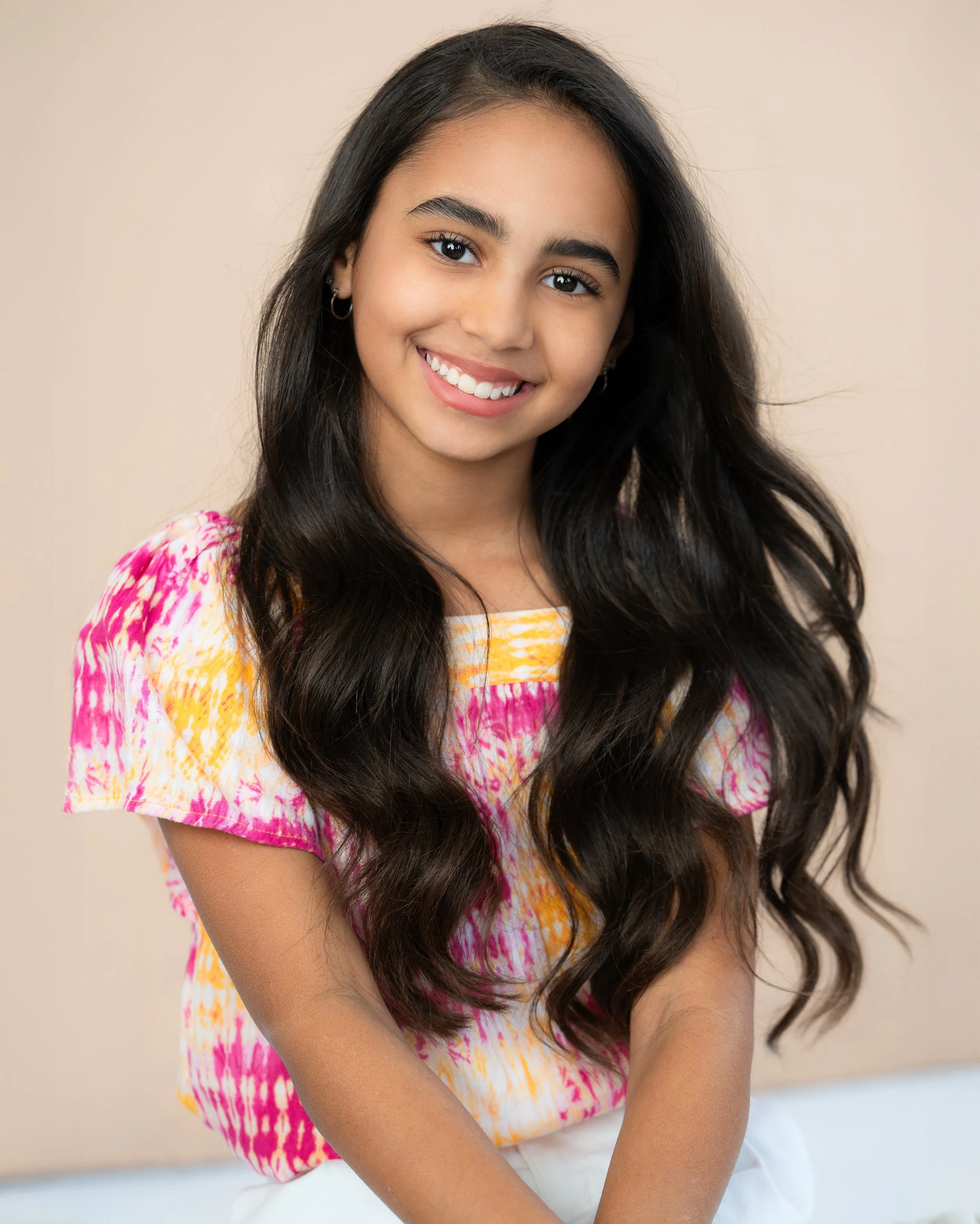A young girl with long dark hair, smiling, wearing a tie-dye pink and yellow shirt and white pants, sitting against a neutral background.