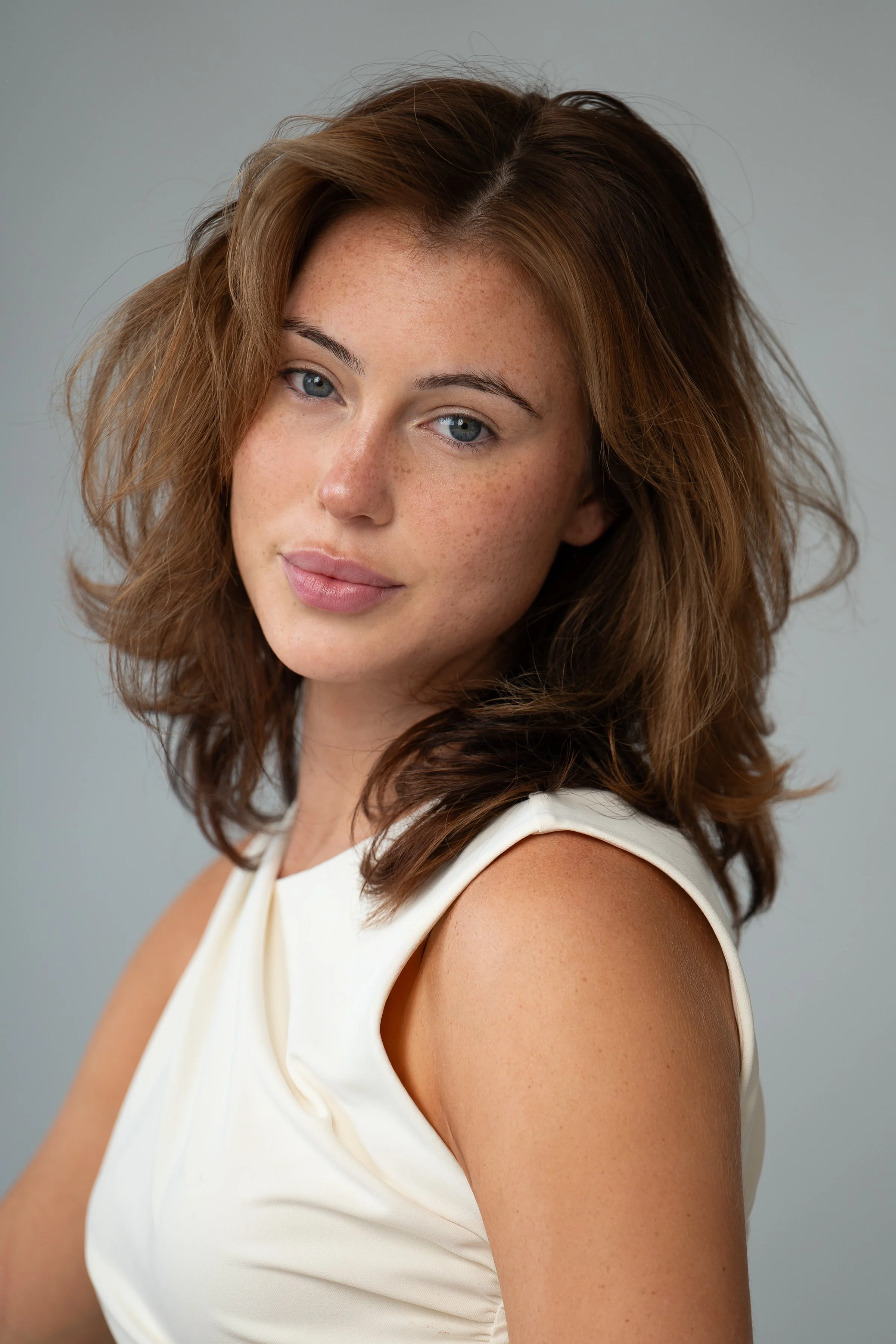 Close-up of a young woman with fair skin, brown wavy hair, light blue eyes, and freckles, wearing a sleeveless white top against a plain gray background.