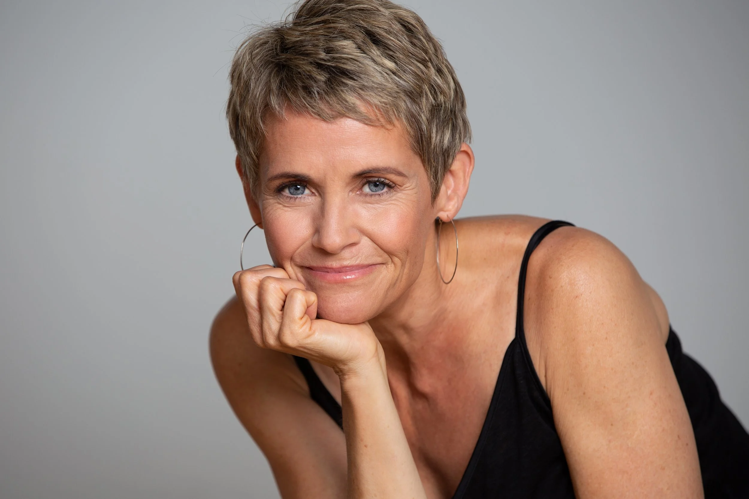 A middle-aged woman with short, blonde hair and blue eyes. She is smiling subtly, resting her chin on her right hand with her fingers curled into a fist. She is wearing silver hoop earrings and a black sleeveless top. The background is plain and ligh