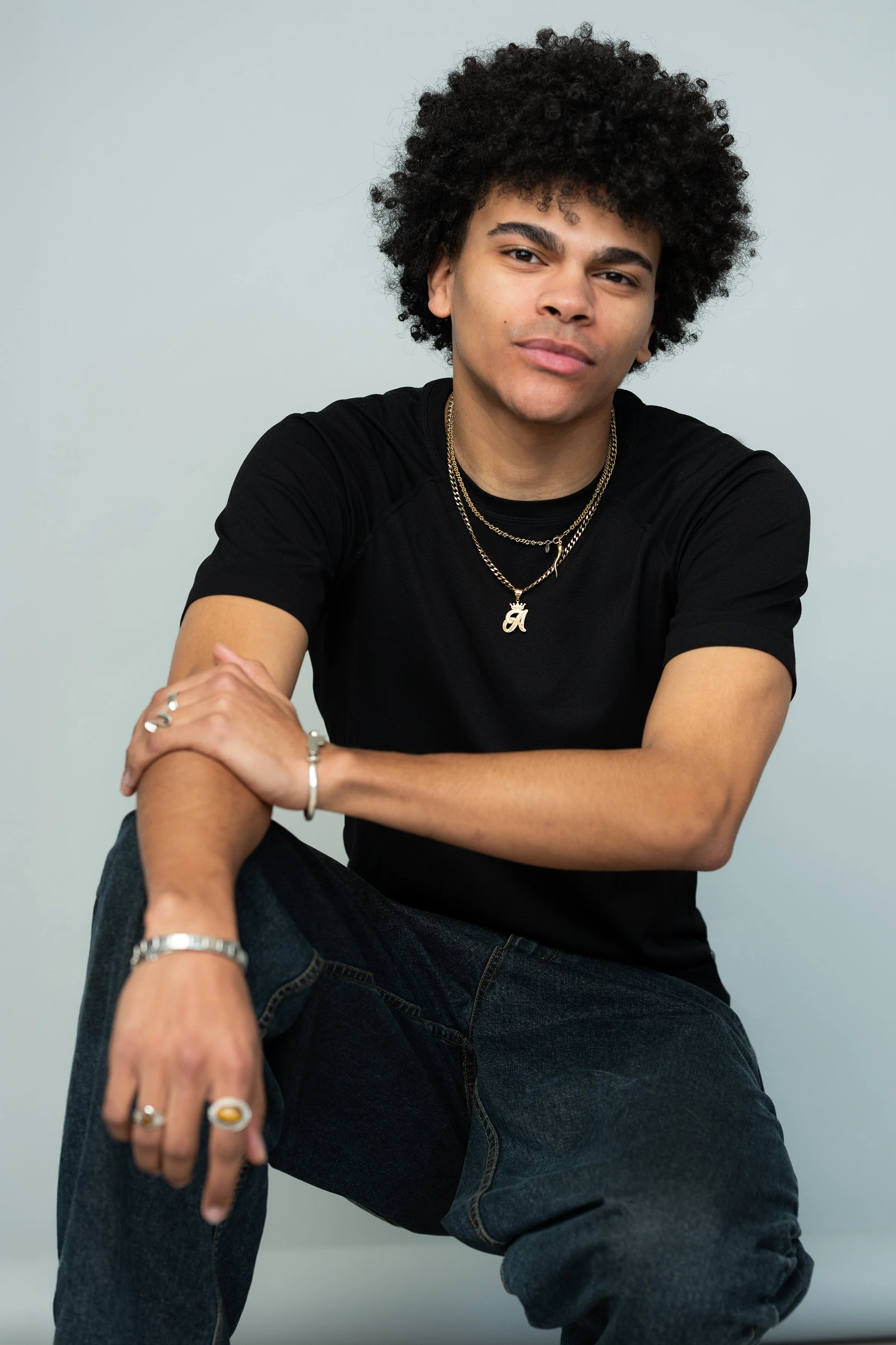 A young man with curly black hair wearing a black T-shirt and layered gold necklaces, sitting casually with one arm resting on his knee and looking at the camera against a plain light gray background.