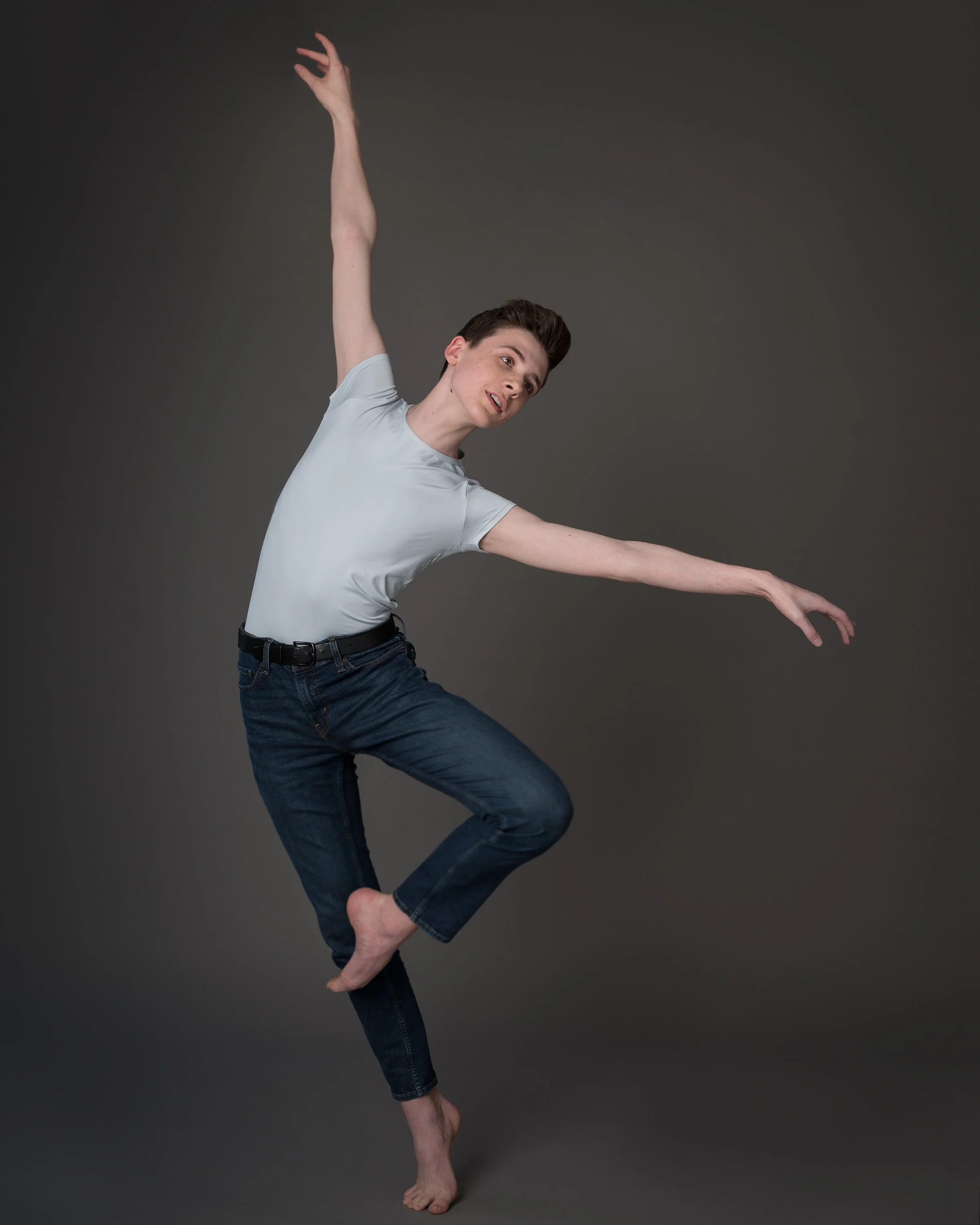 A person with short brown hair and light skin is standing on one foot while balancing with arms extended and bent, against a dark gray background.