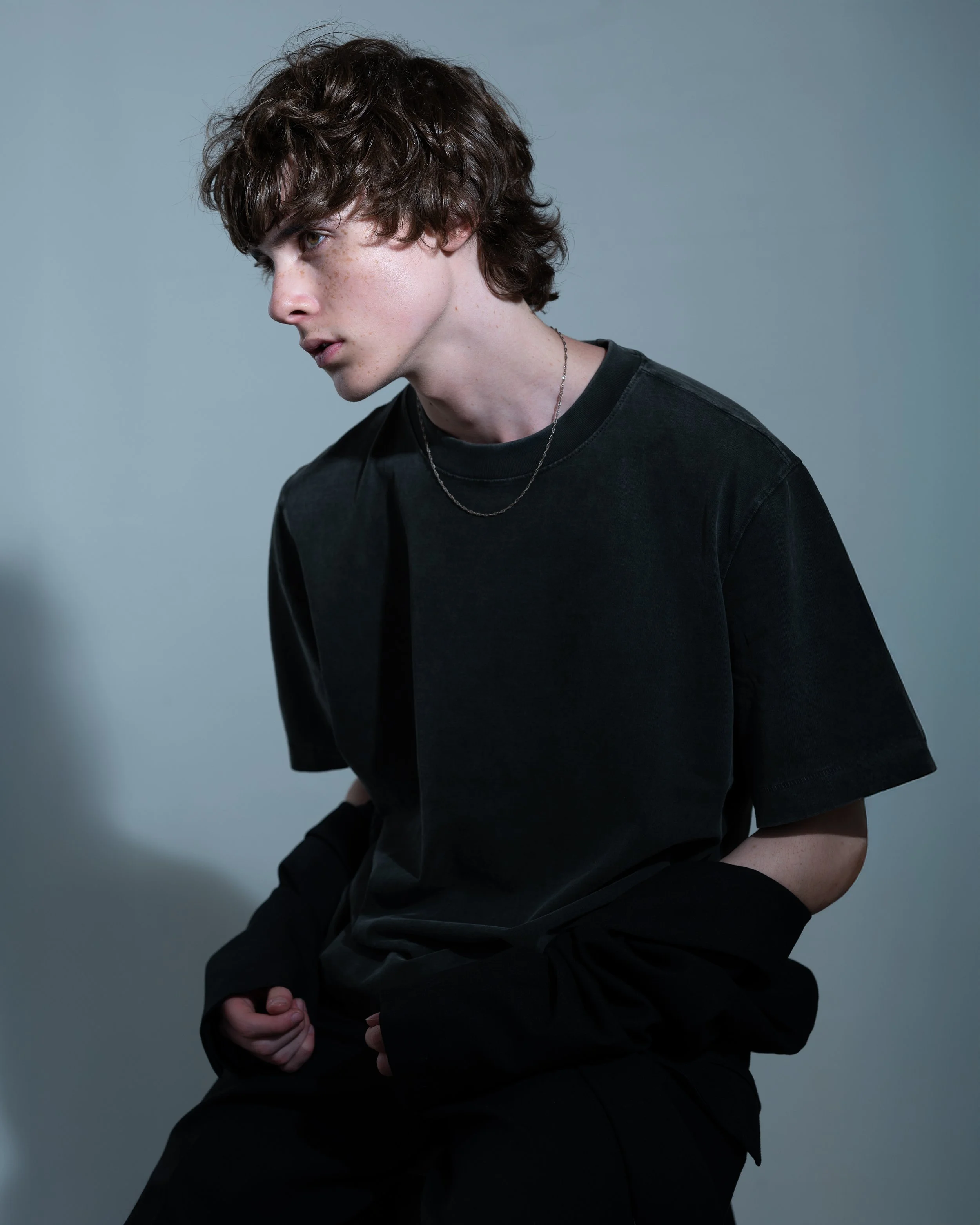 A young person with curly brown hair, light skin, and freckles wearing a black t-shirt and a silver chain necklace, posed against a plain gray background.