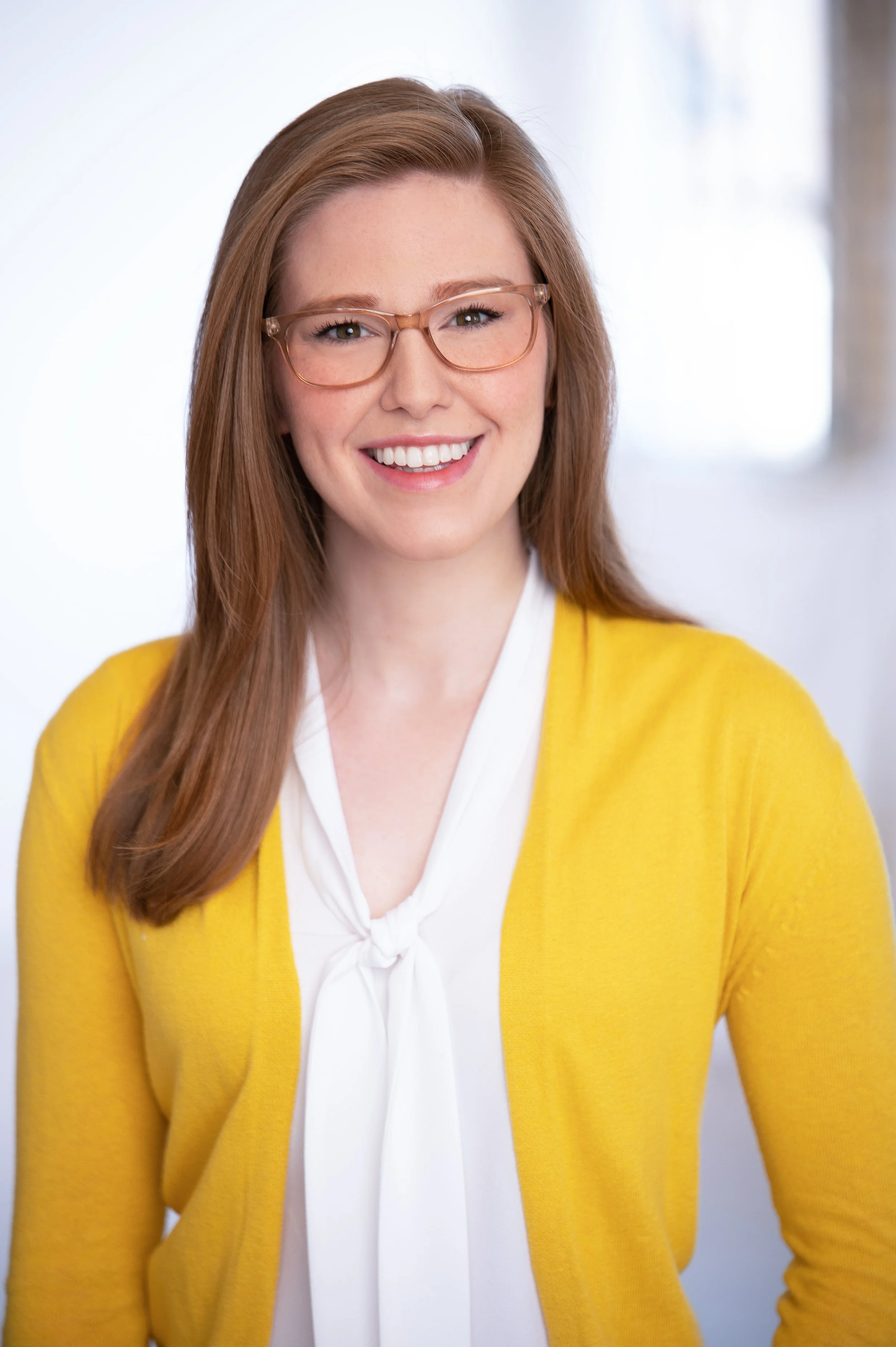 Close-up of a woman with long red hair, wearing glasses, a white blouse, and a yellow cardigan, smiling at the camera.