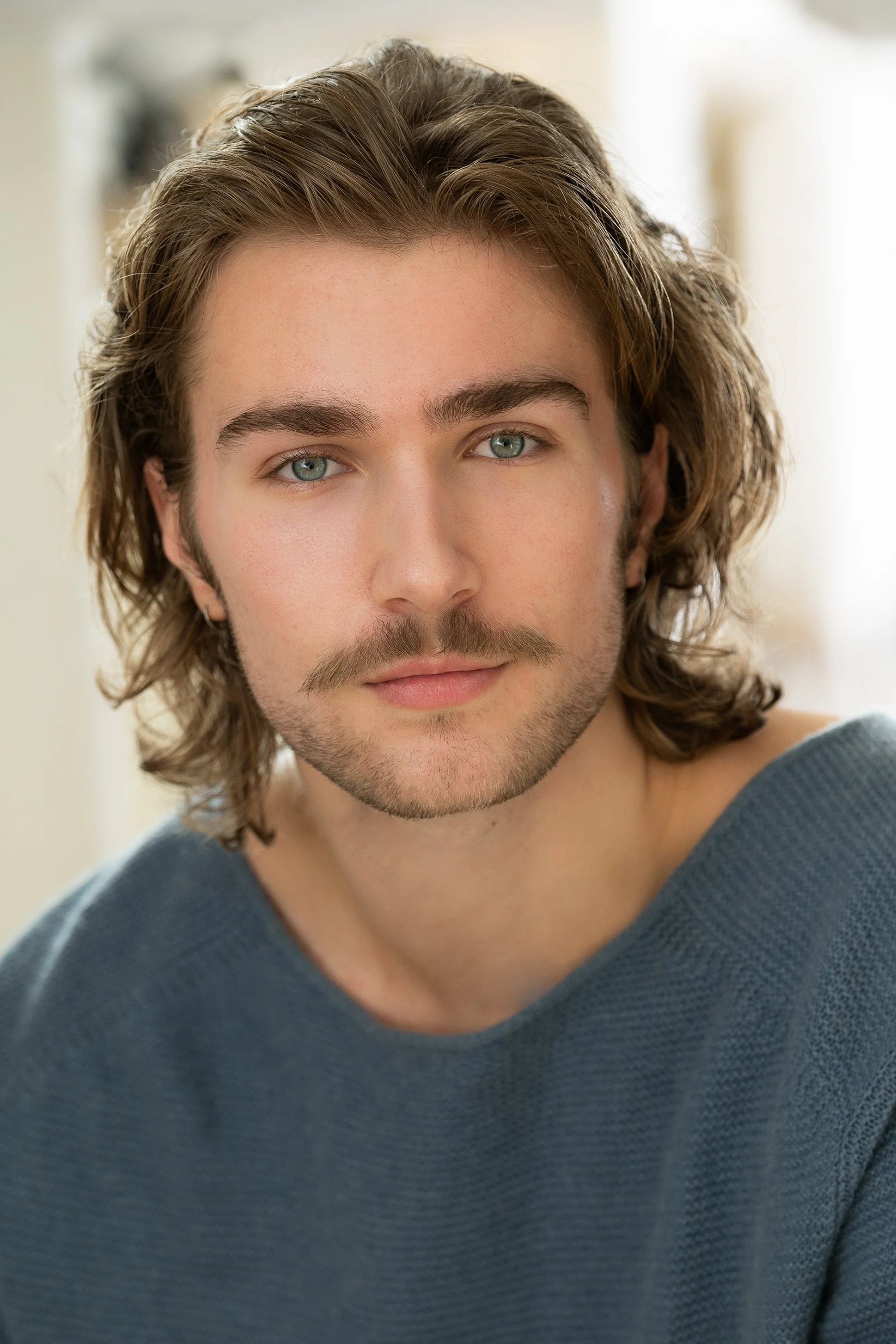 Close-up of a young man with wavy brown hair, blue eyes, and light facial hair, wearing a blue sweater.