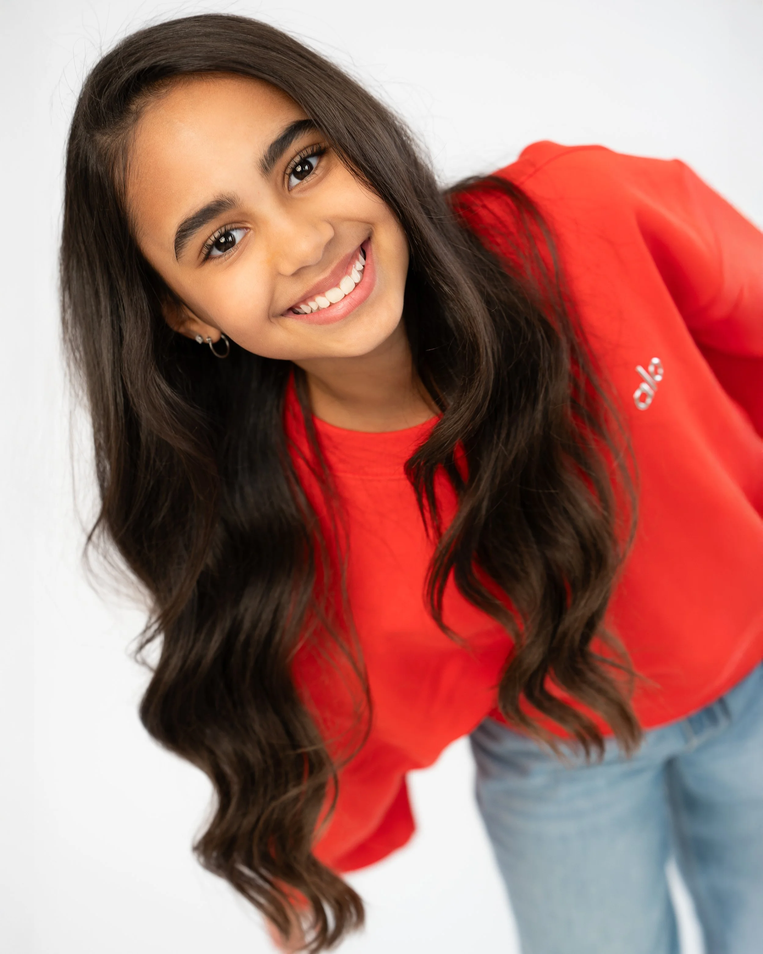 Close-up of a young woman with long dark hair, smiling, wearing a red shirt and light blue jeans, against a white background.
