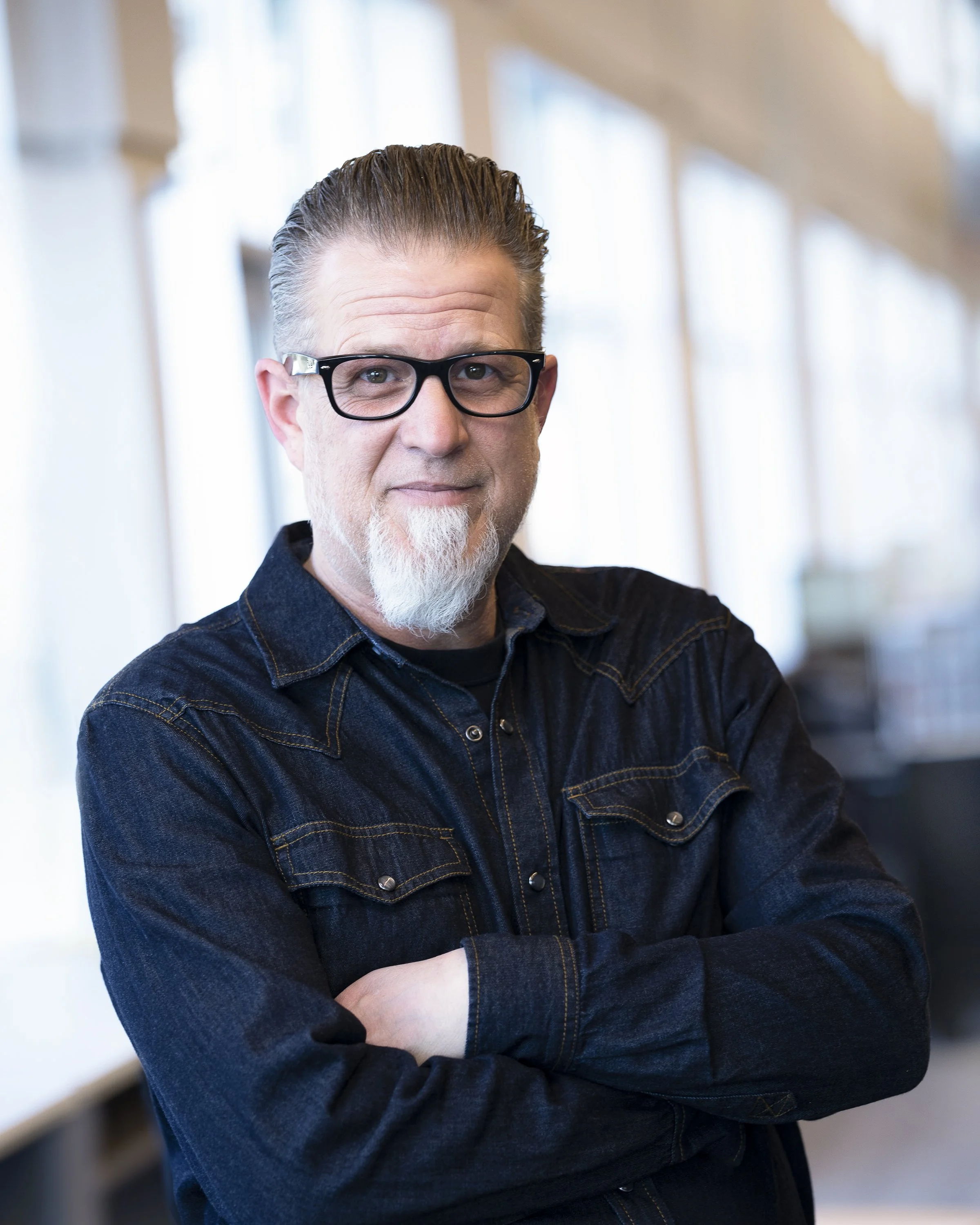 A man with gray hair, a beard, and glasses, wearing a dark denim shirt, smiling and standing indoors with natural light in the background.