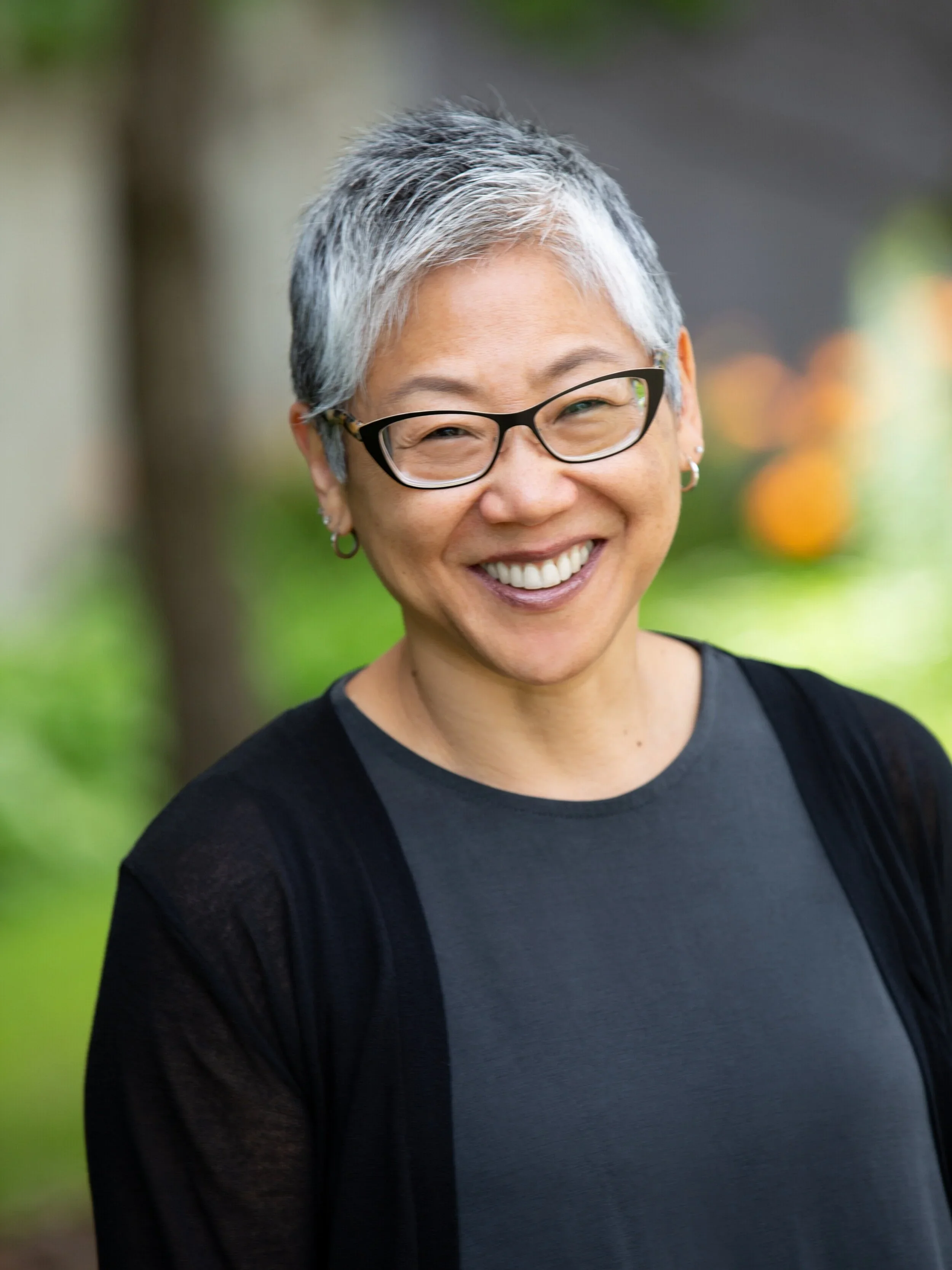 Smiling woman with short gray hair, black glasses, earrings, wearing a black top with gray accents, outdoors with blurred green and orange background.