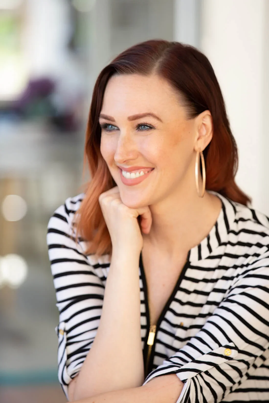 A woman with red hair, blue eyes, and hoop earrings wearing a black and white striped top, smiling with her chin resting on her hand.