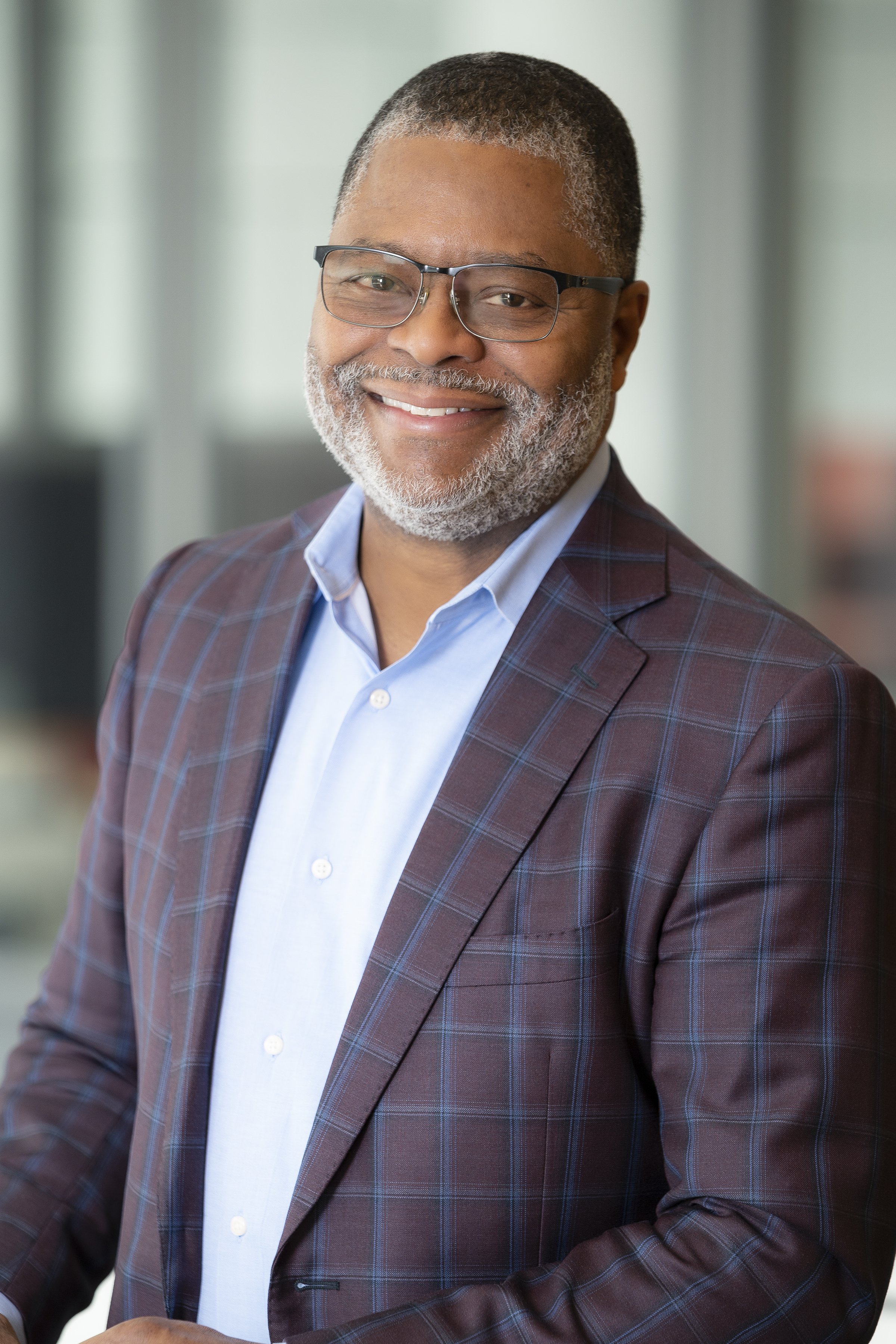 A middle-aged man with a beard and glasses, wearing a brown plaid blazer and a light blue shirt, smiling indoors with a blurred background.