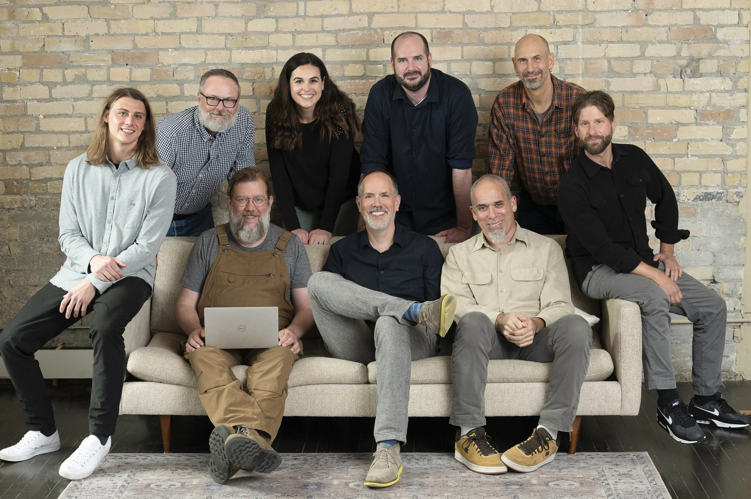 A diverse group of ten people posing in front of an exposed brick wall. Some are sitting on a beige sofa, others are standing or kneeling behind it. They are smiling and casually dressed, with one person using a laptop.