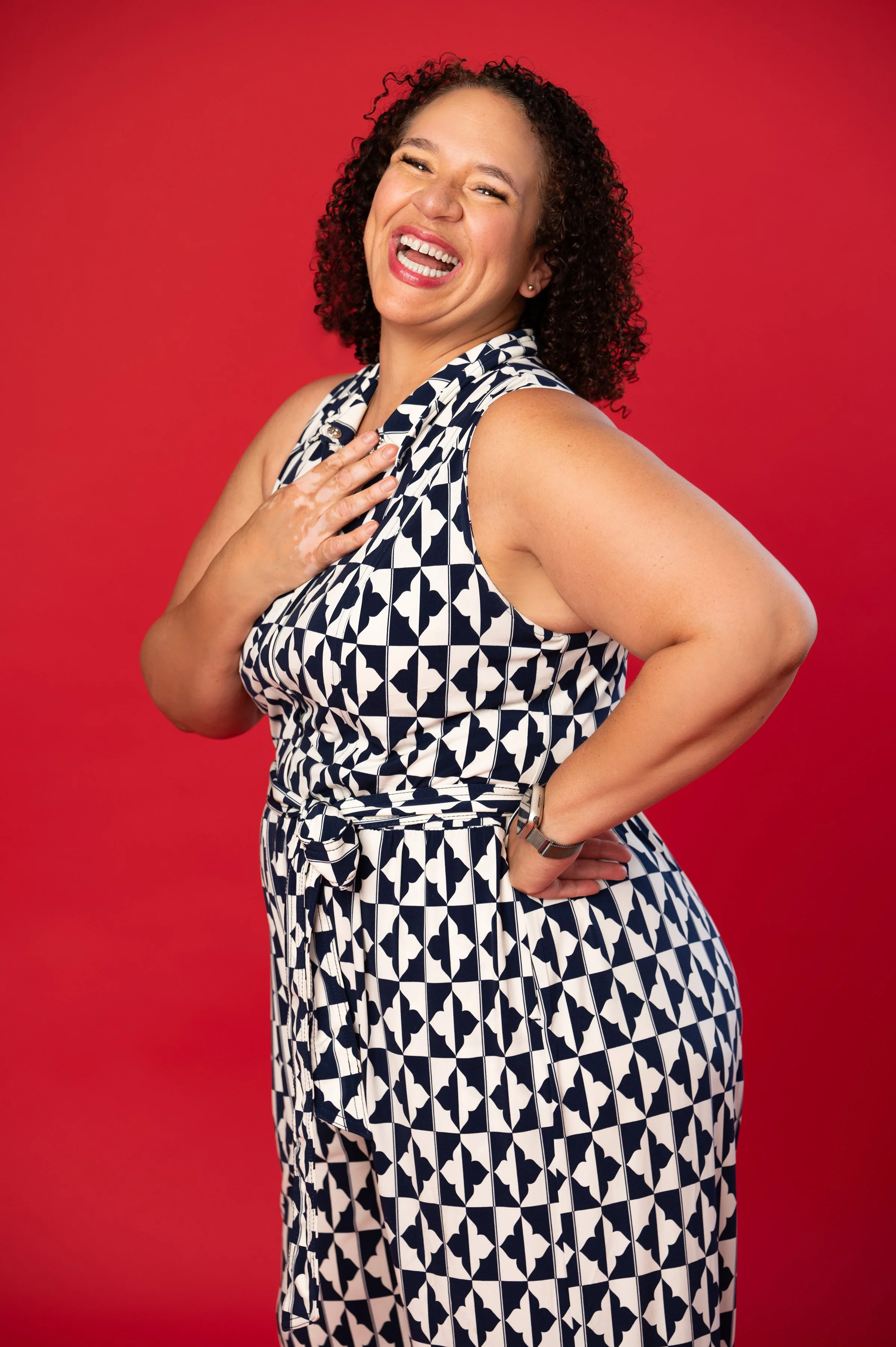A woman with curly hair smiling and showing her teeth, wearing a blue and white patterned dress, standing against a red background.