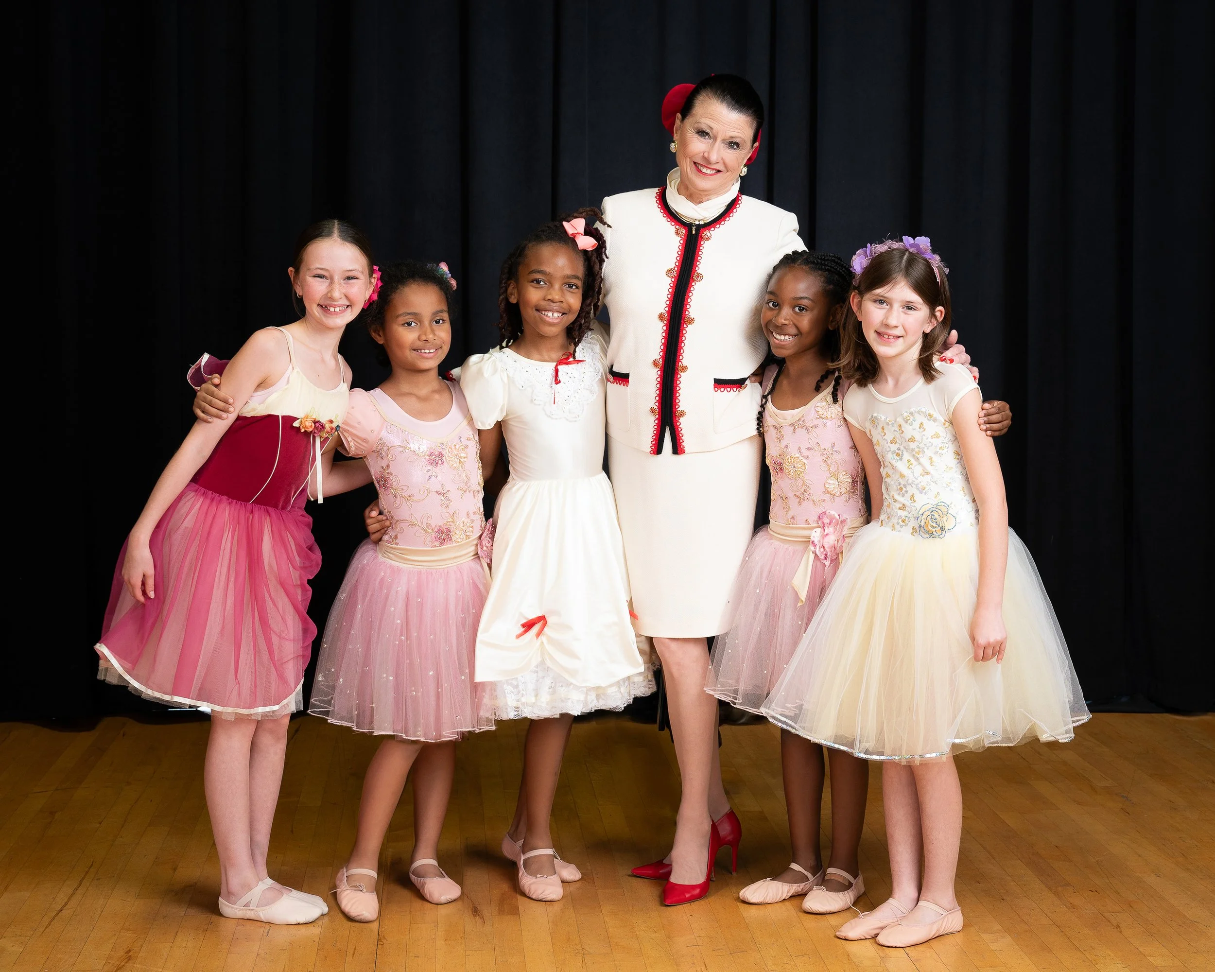 Ballet instructor with five young girls in ballet costumes on stage with black curtains.