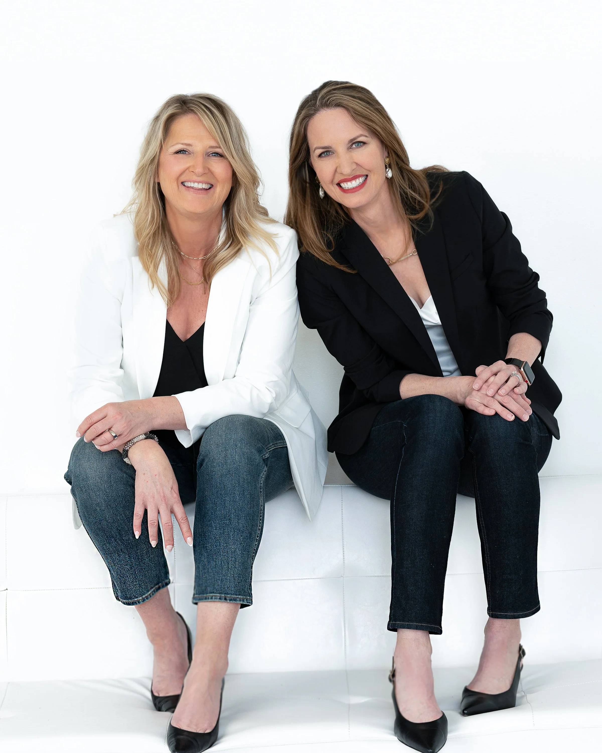 Two women sitting on a white couch against a white background, smiling. One woman is wearing a white blazer over a black top, jeans, and black heels. The other woman is wearing a black blazer over a white shirt, jeans, and black heels. Both have jewe