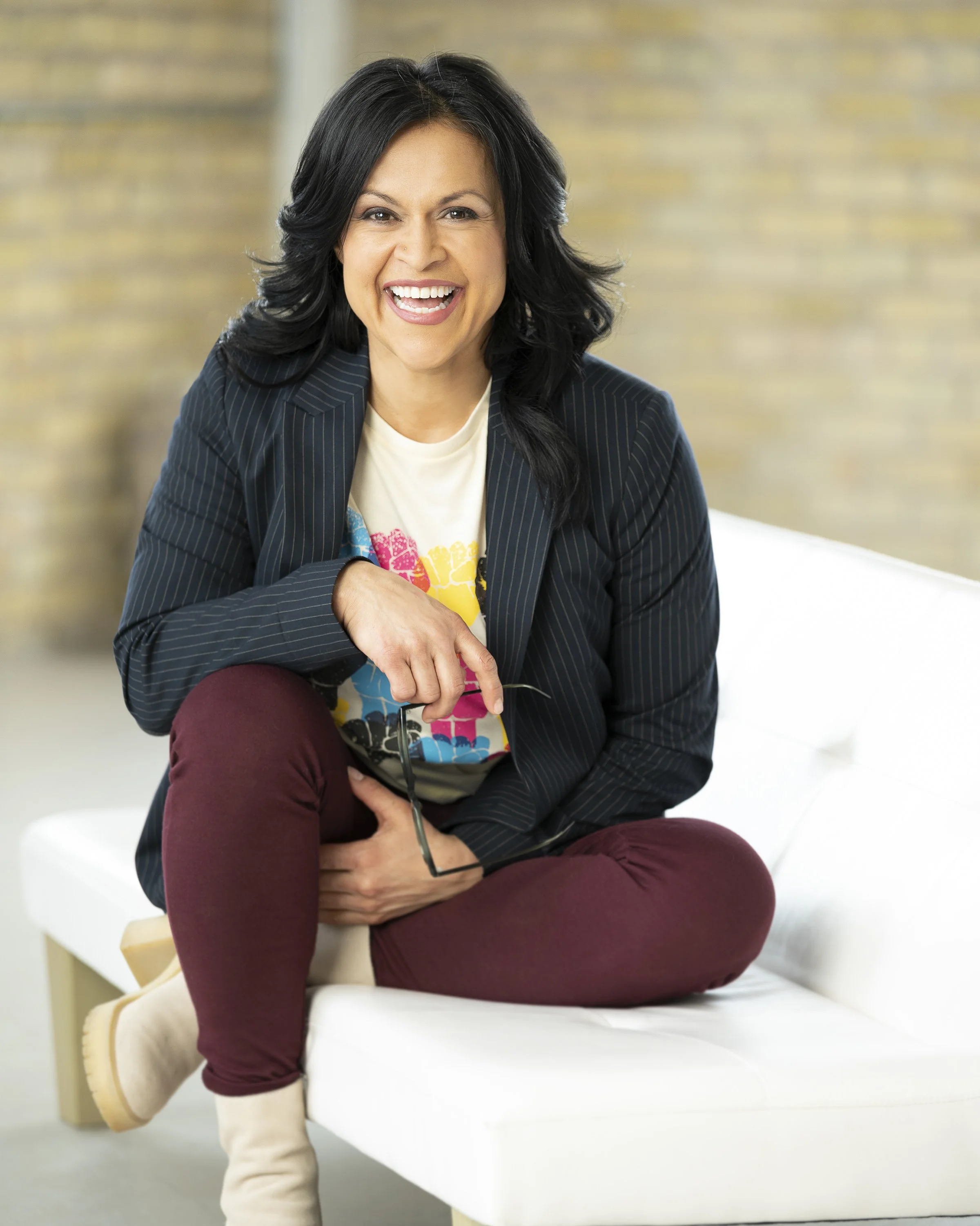 Woman with black hair, wearing a dark pinstripe blazer and a white shirt with colorful pattern, sitting on a white bench, laughing, holding glasses in her right hand, with legs crossed, beige ankle boots, against blurred background.