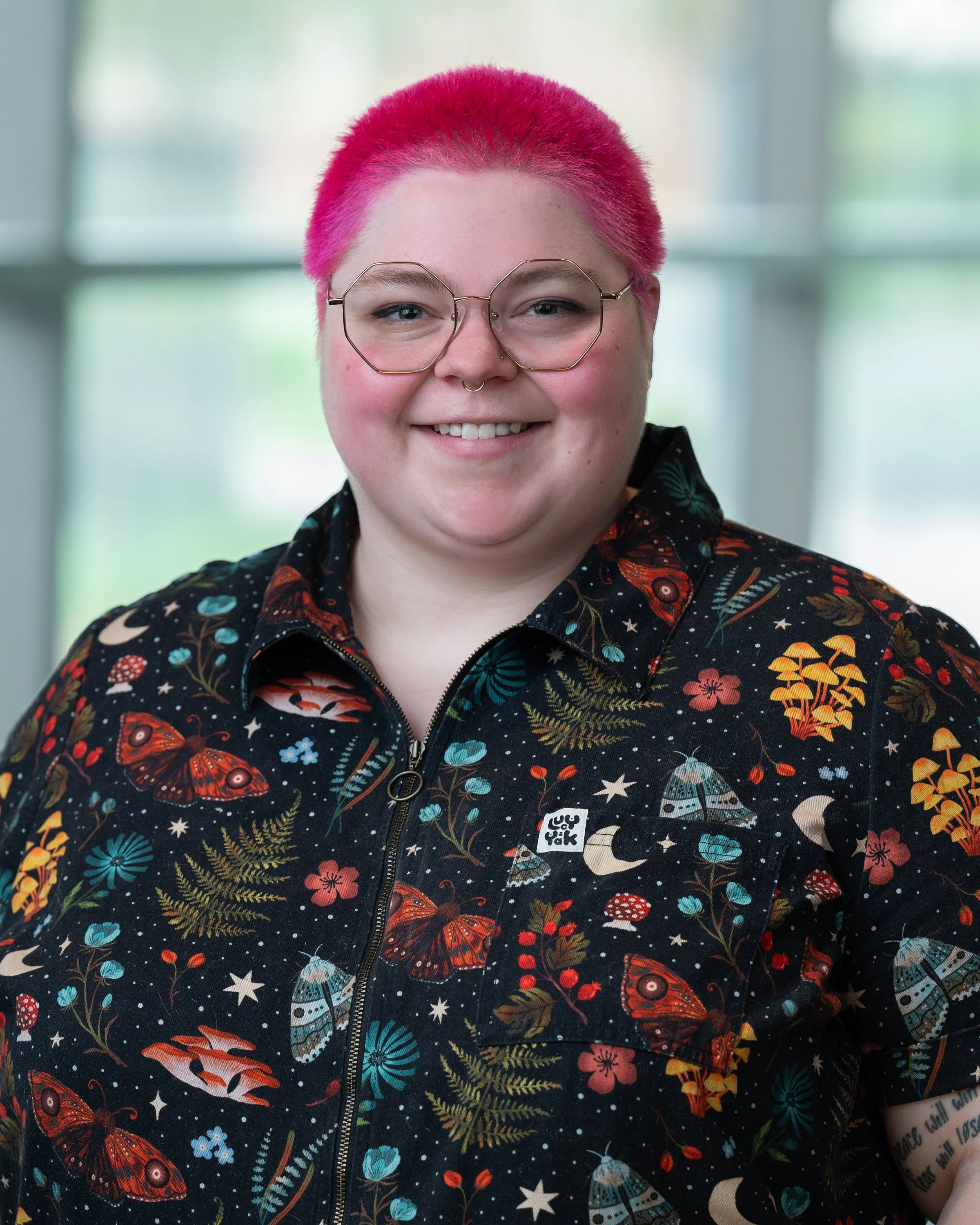 A person with pink and blonde hair, wearing round glasses and a black shirt with a colorful space-themed and nature print, smiling indoors in front of windows.