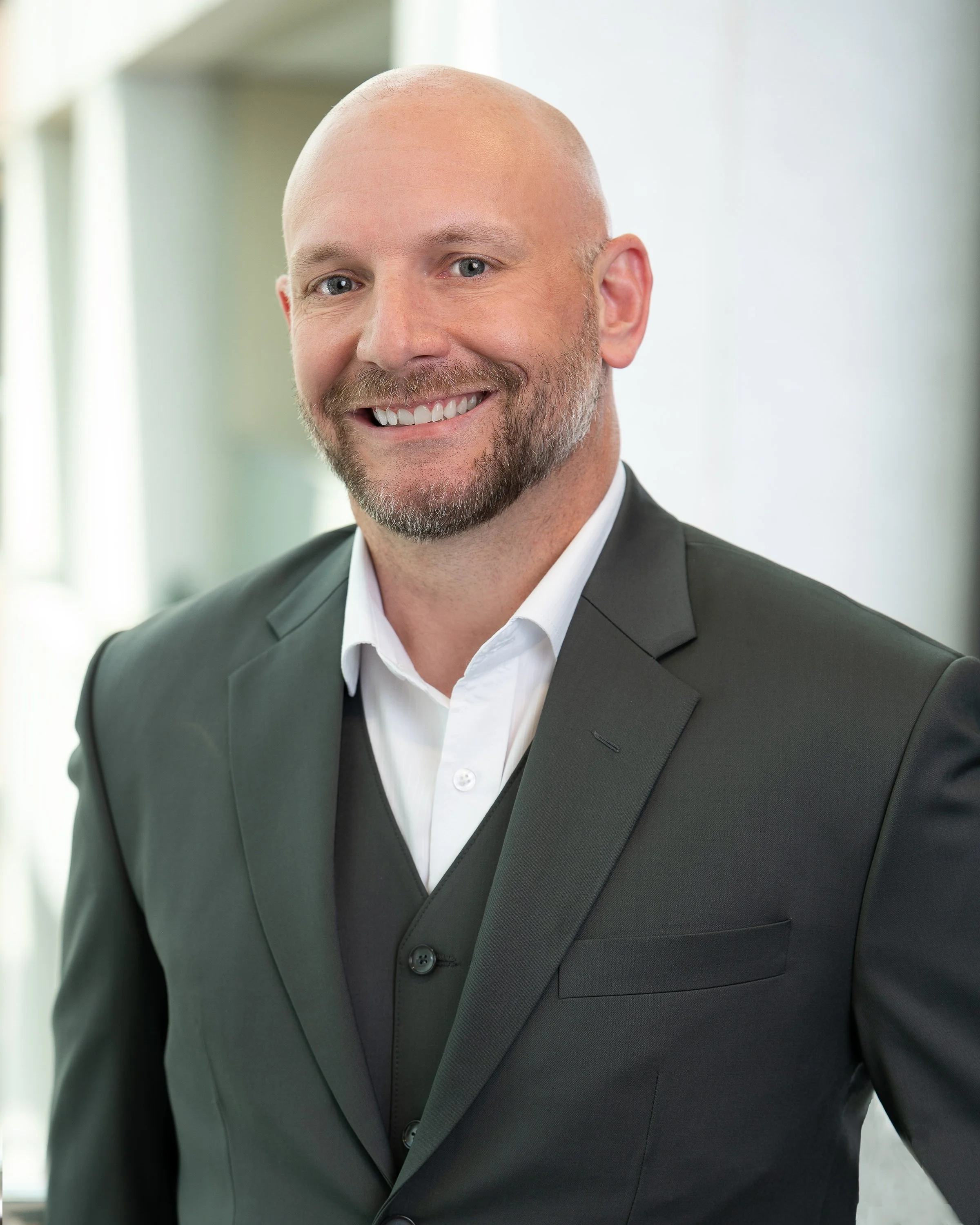 A man with a bald head and gray beard smiling, wearing a dark suit and white dress shirt, in an office environment.