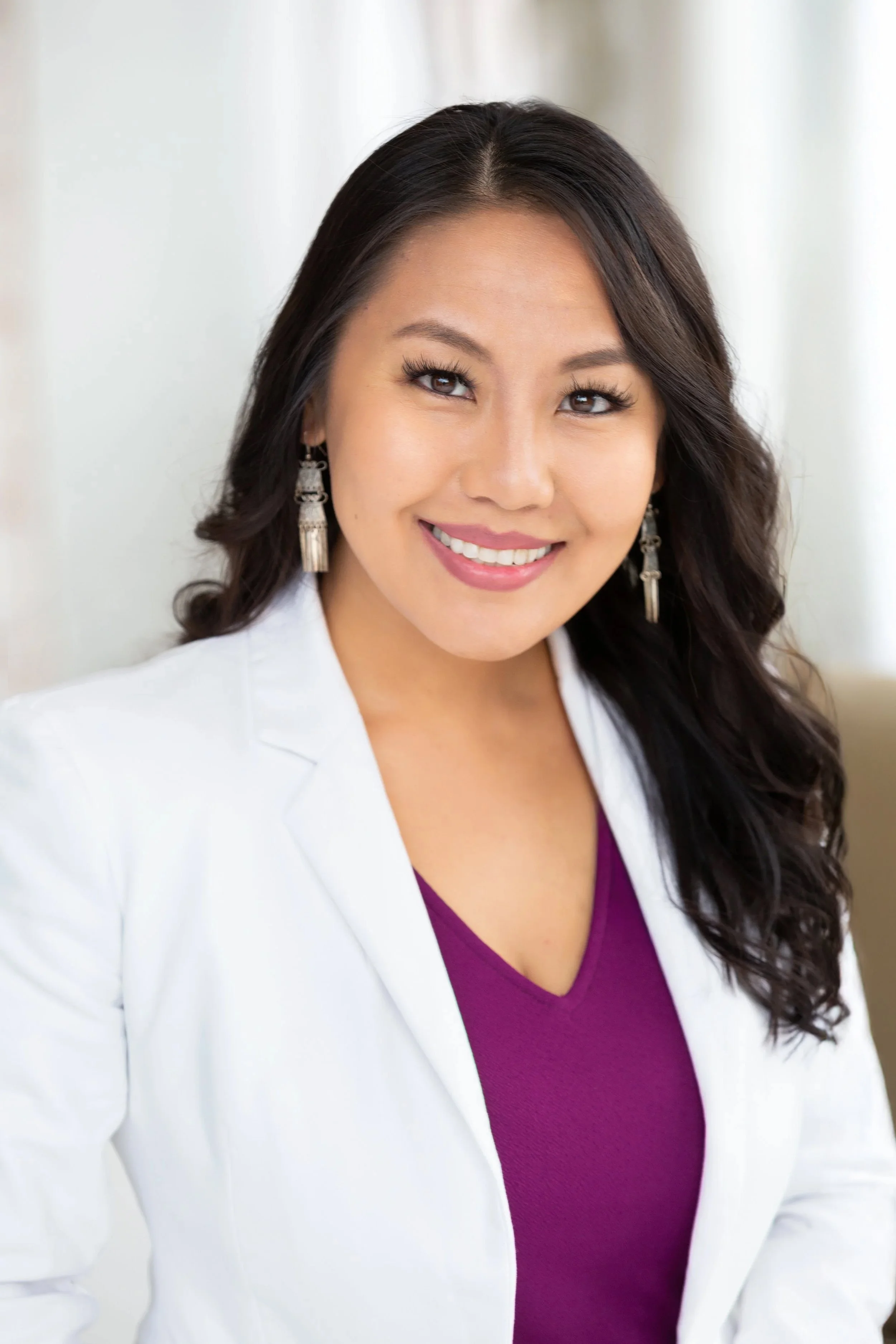 A woman with long dark hair and earrings, smiling, wearing a purple top and a white blazer.