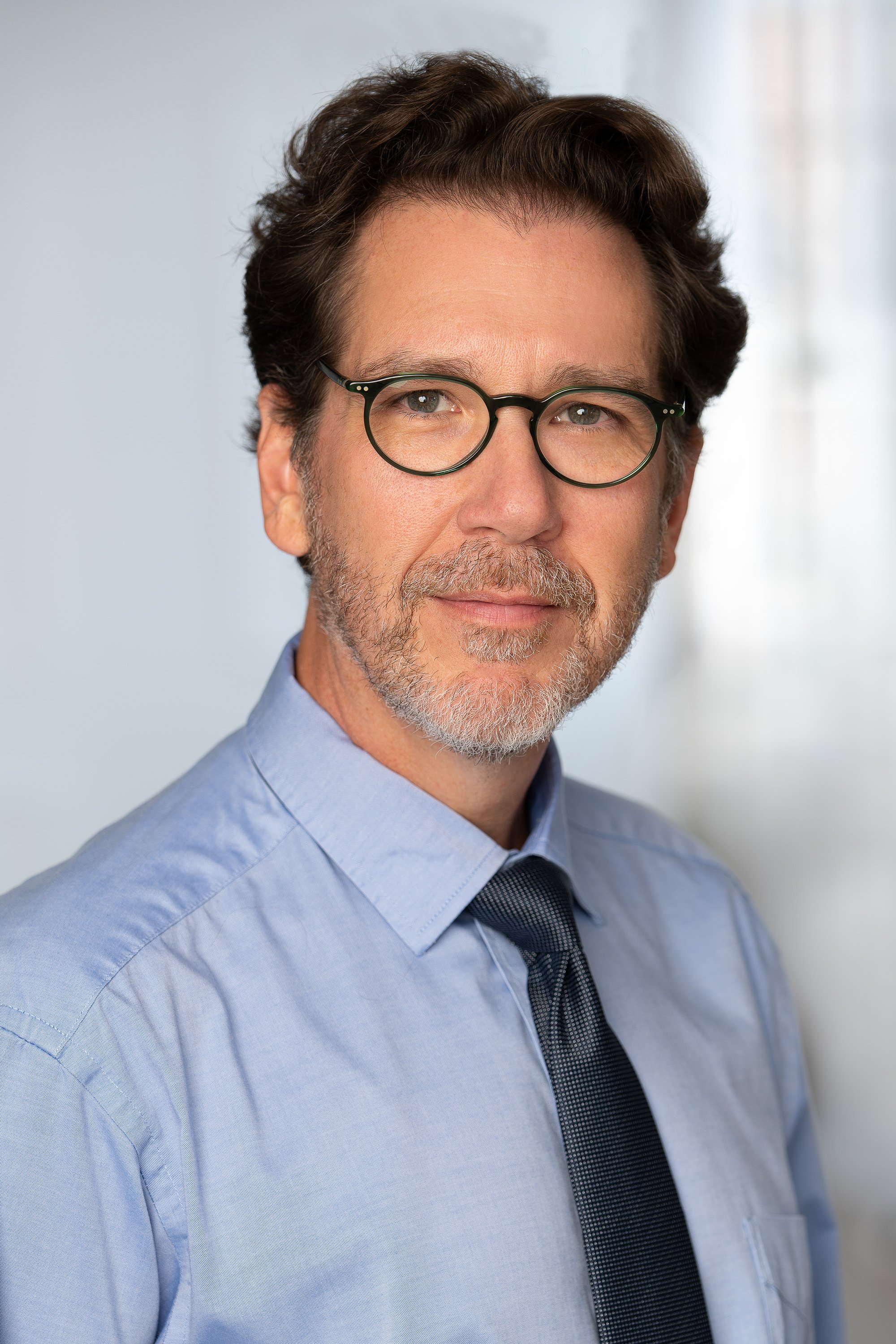 Close-up portrait of a middle-aged man with brown hair, glasses, and a beard, wearing a light blue dress shirt and a dark tie, standing against a light background.
