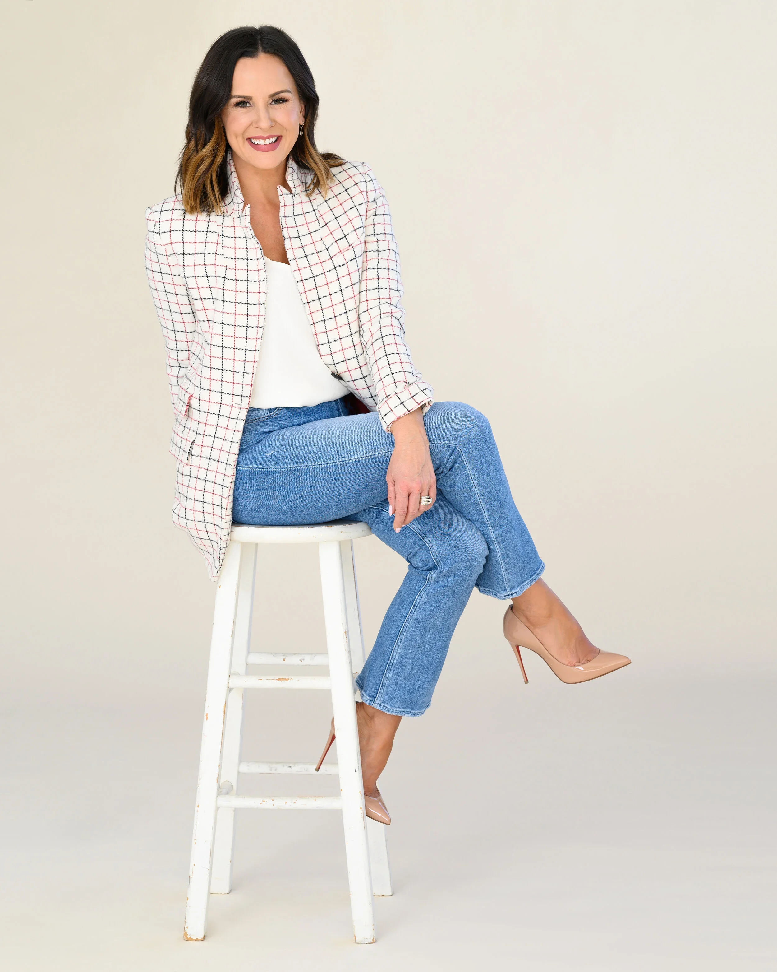 Woman smiling, dressed in a checked blazer and jeans, sitting on a white stool against a minimalist beige background, with her left leg crossed over her right, wearing nude high heels.
