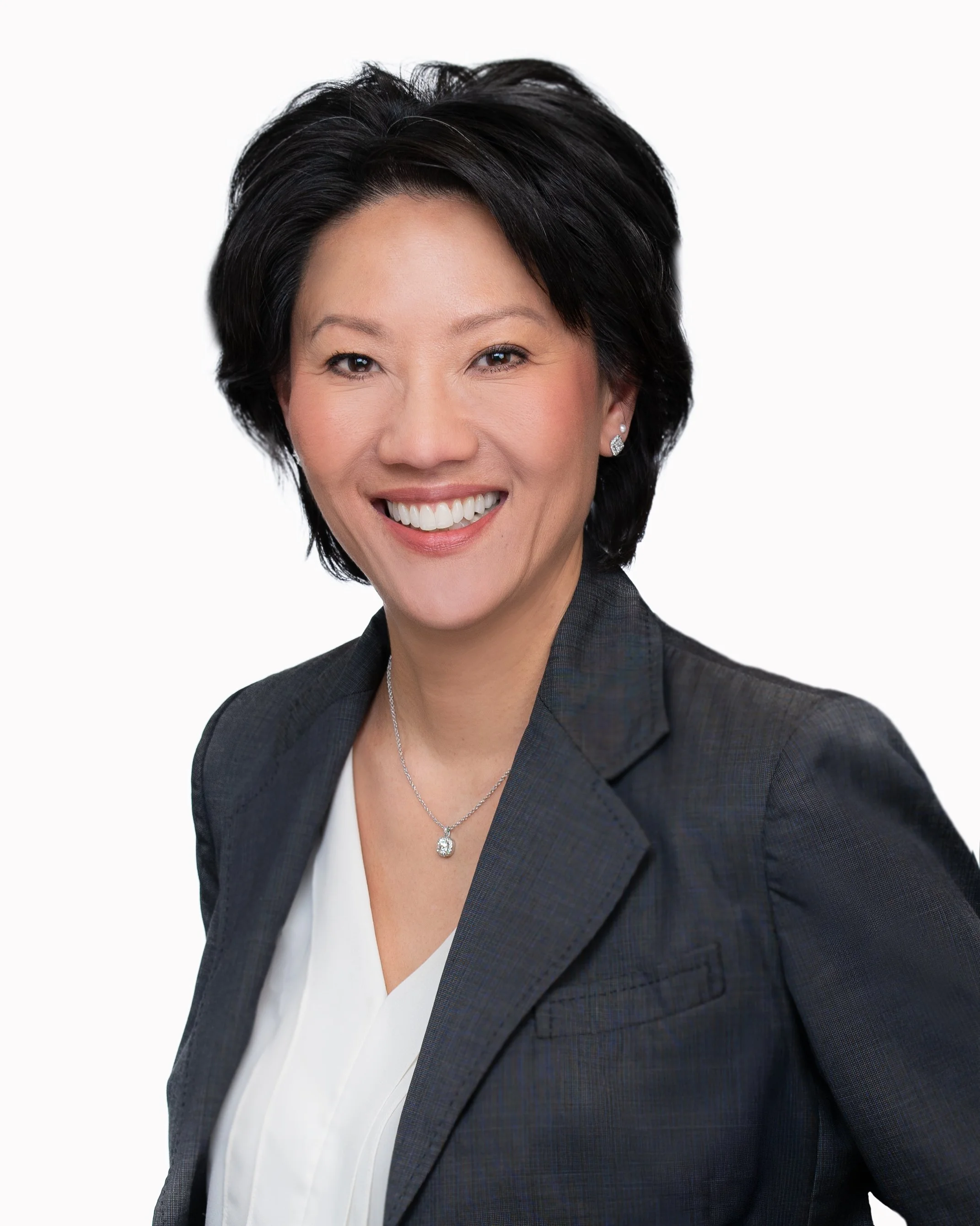 A woman with short black hair, smiling, wearing a dark gray blazer, a white blouse, a necklace, and earrings, against a plain white background.
