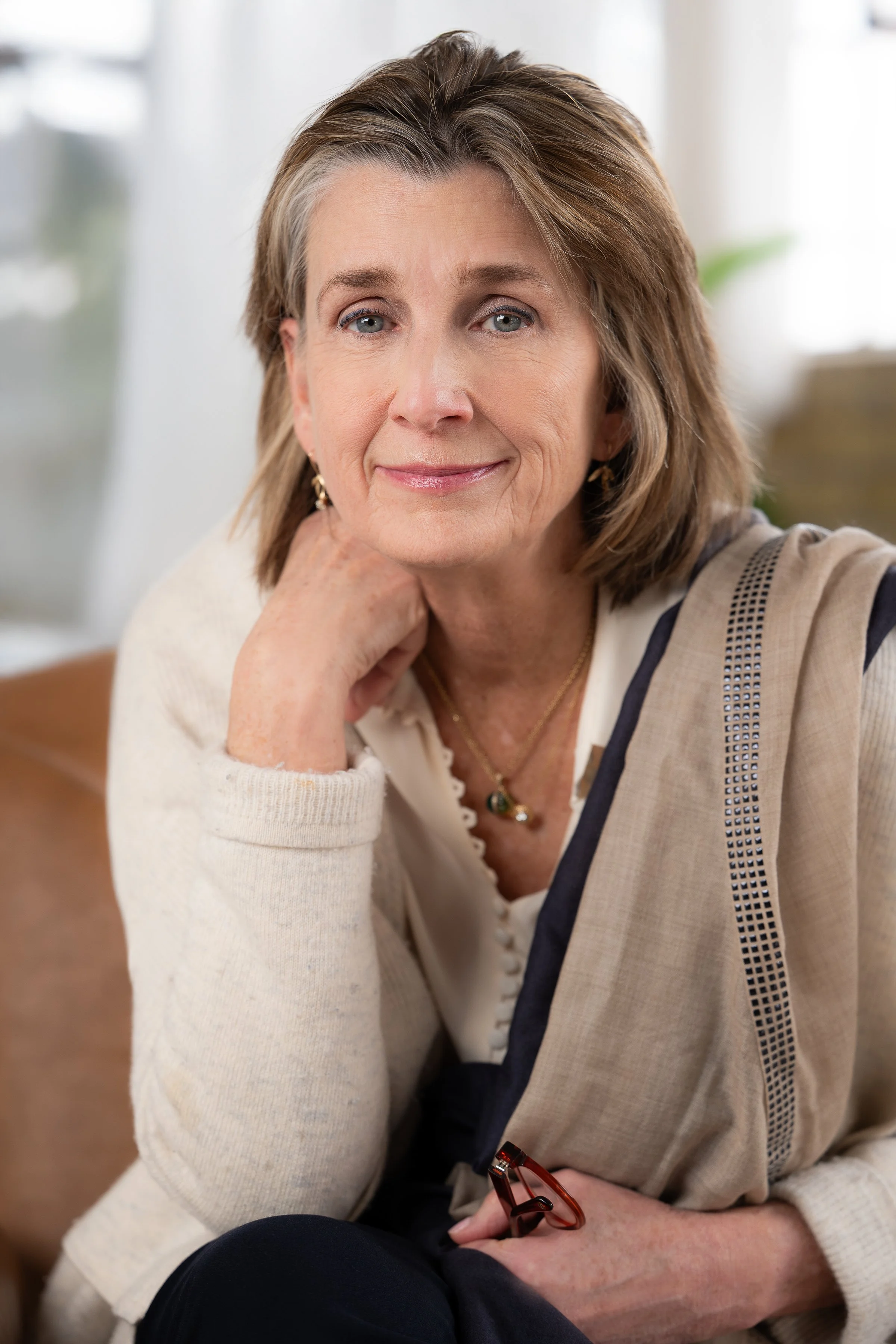 A middle-aged woman with short brown hair, blue eyes, and light skin, smiling softly at the camera, sitting indoors with a blurred background, wearing a cream sweater with navy accents, gold earrings, and a gold necklace.