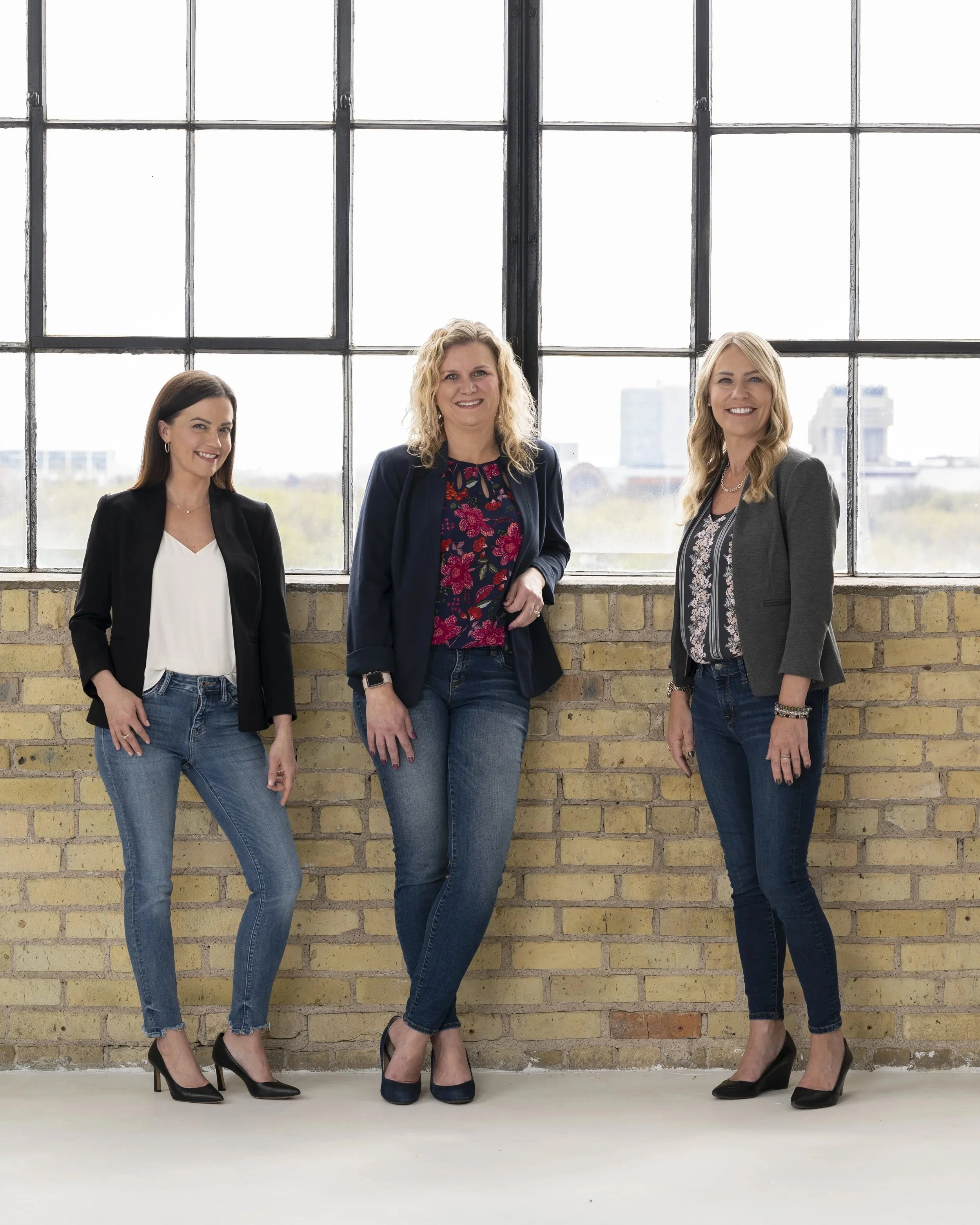 Three women standing in front of large window with cityscape background, all dressed in business casual attire, smiling.